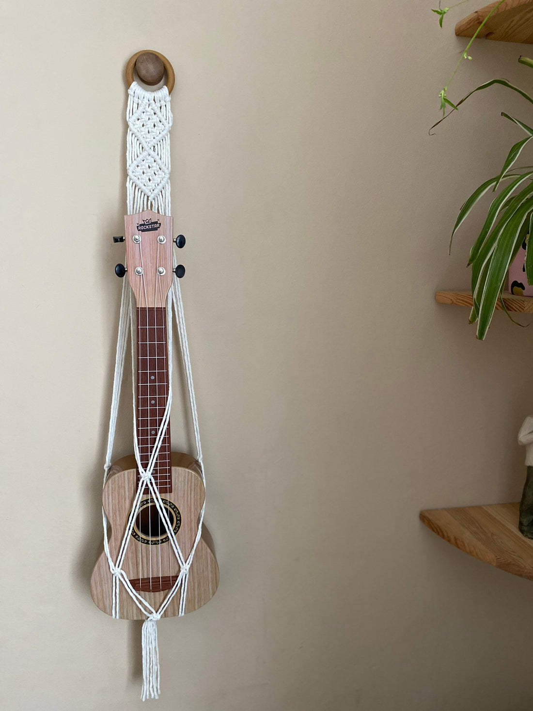 A light brown ukulele sits supported inside a macrame ukulele hanger, made with natural yarn and wooden ring, against a beige wall.