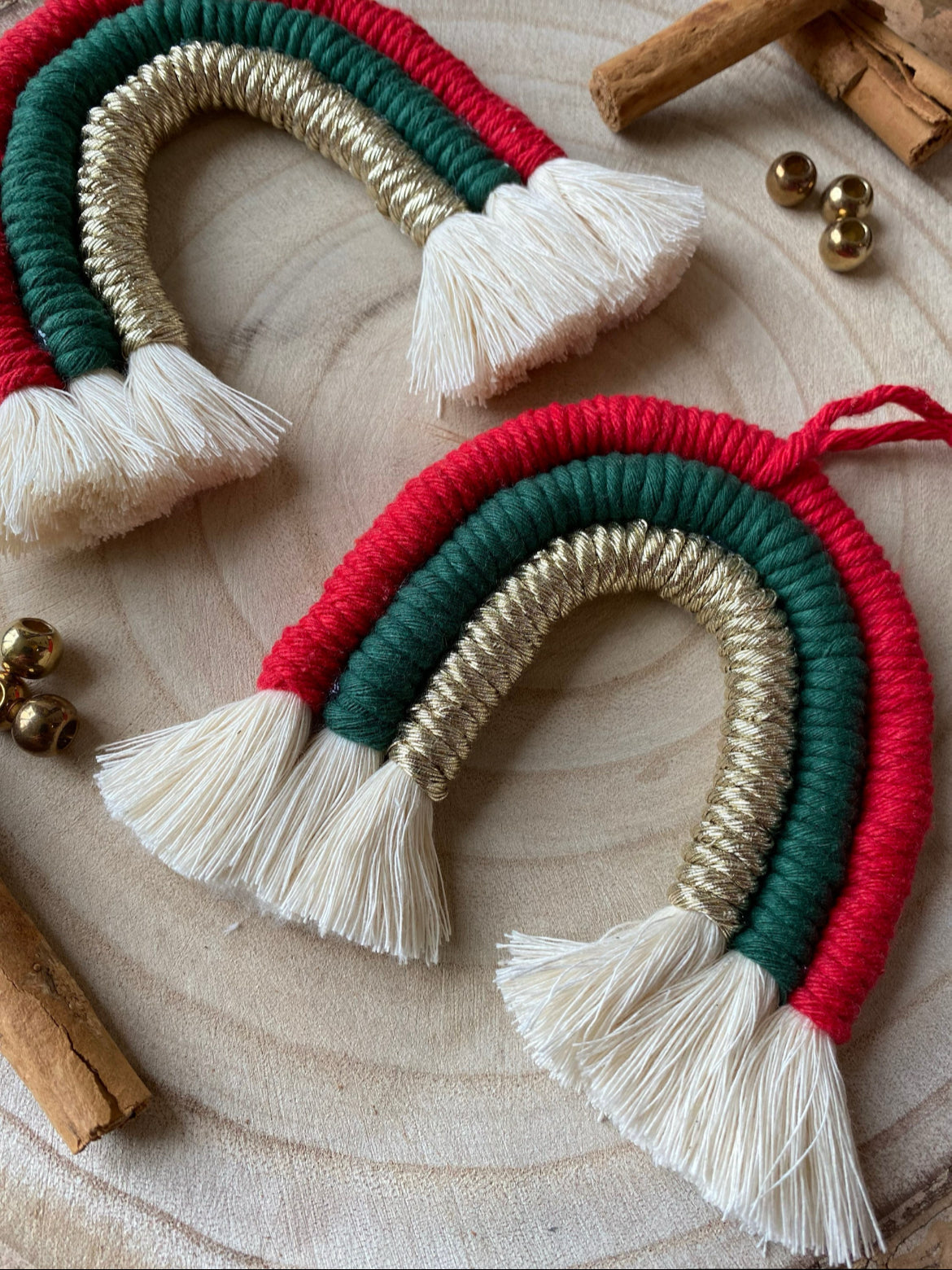 Two macrame rainbows made with red, green and gold yarns, lay on a wooden surface surrounded by small gold beads and cinnamon sticks.
