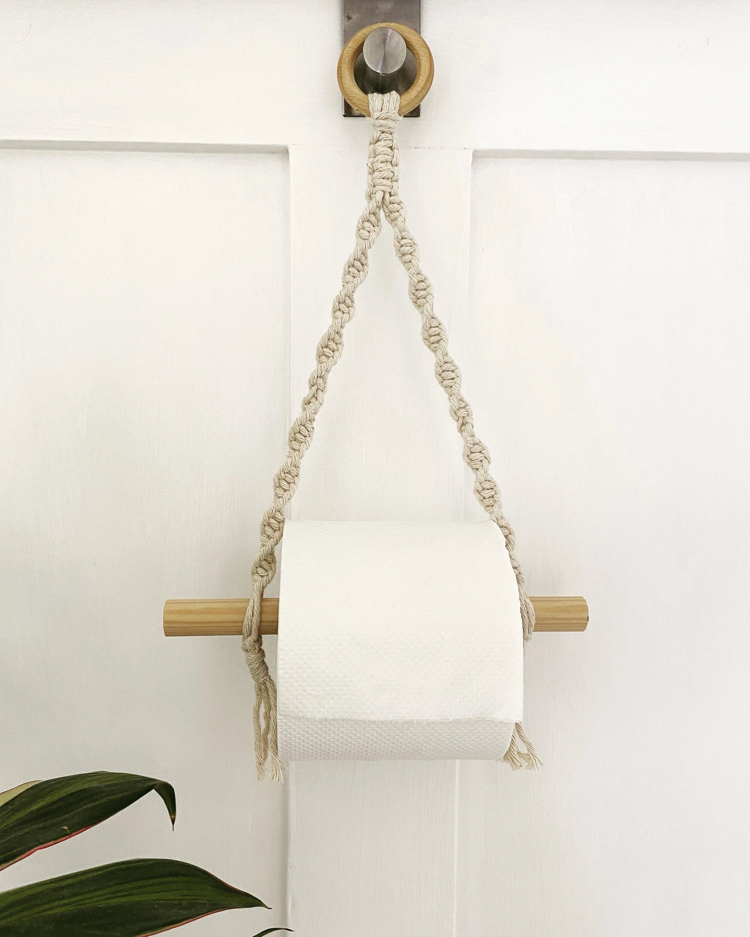 Macrame toilet roll holder made with natural cotton and a wooden hanging ring, holding a white toilet roll against the back of a white door.