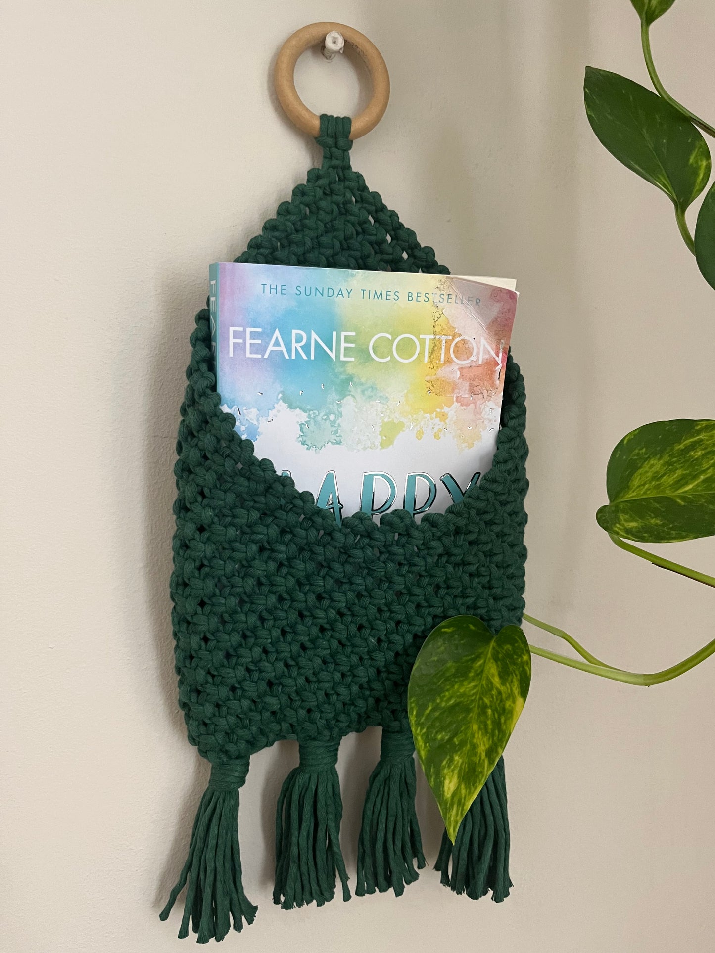 A green, knitted wall pocket crafted from recycled cotton decor hangs from a wooden ring, holding a book titled "Happy." The Macra-Made-With-Love Bedside storage pouch book sleeve has tassels at the bottom, and a pothos plant with heart-shaped leaves partially wraps around the eco-friendly bedroom storage.