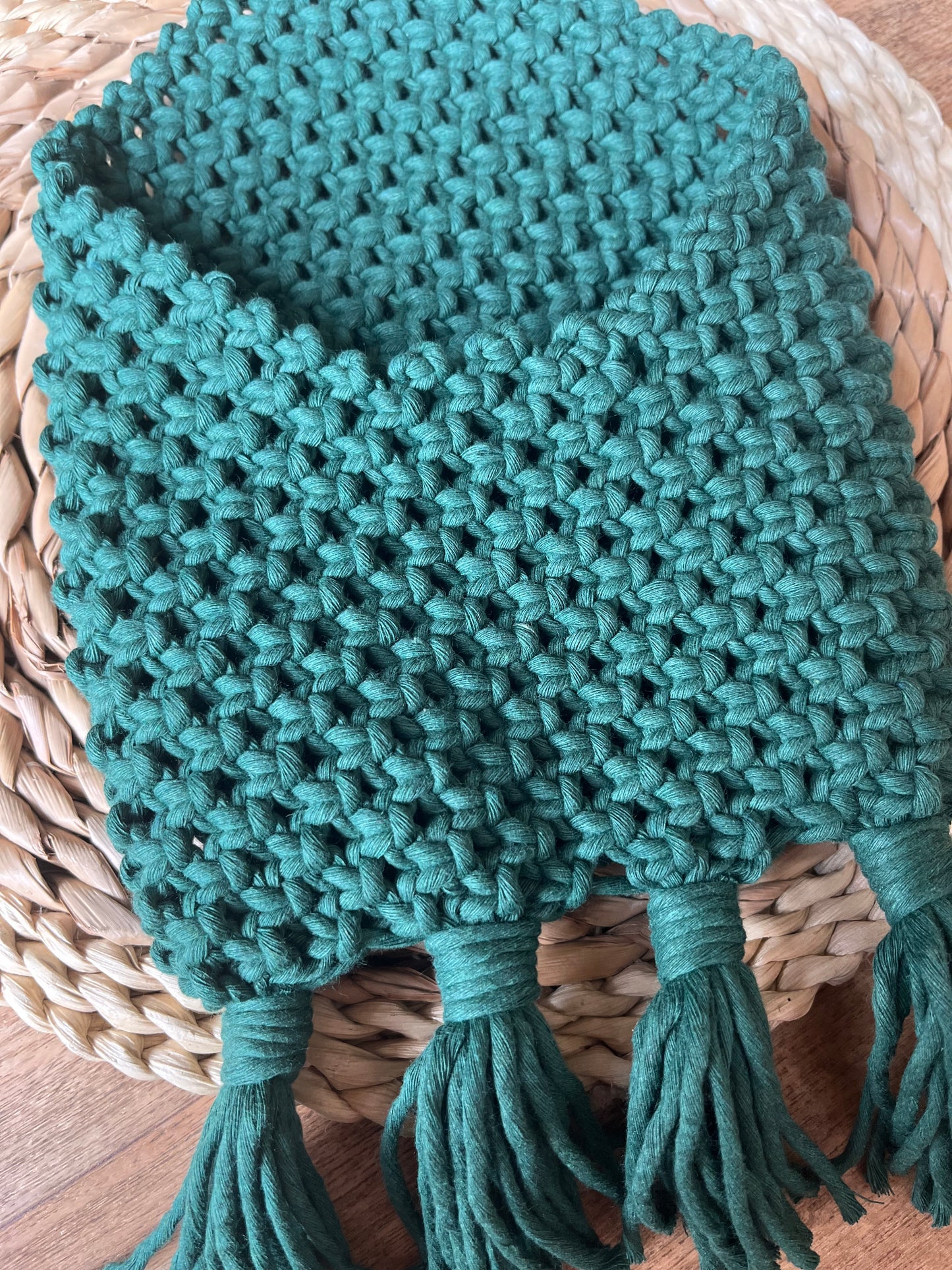 A hand-knitted, green scarf with a chunky texture is placed neatly on a woven straw surface, paired beautifully with a Macra-Made-With-Love Bedside storage pouch book sleeve. The scarf has several tassels hanging from the bottom edge, adding to its cozy and handmade appearance.