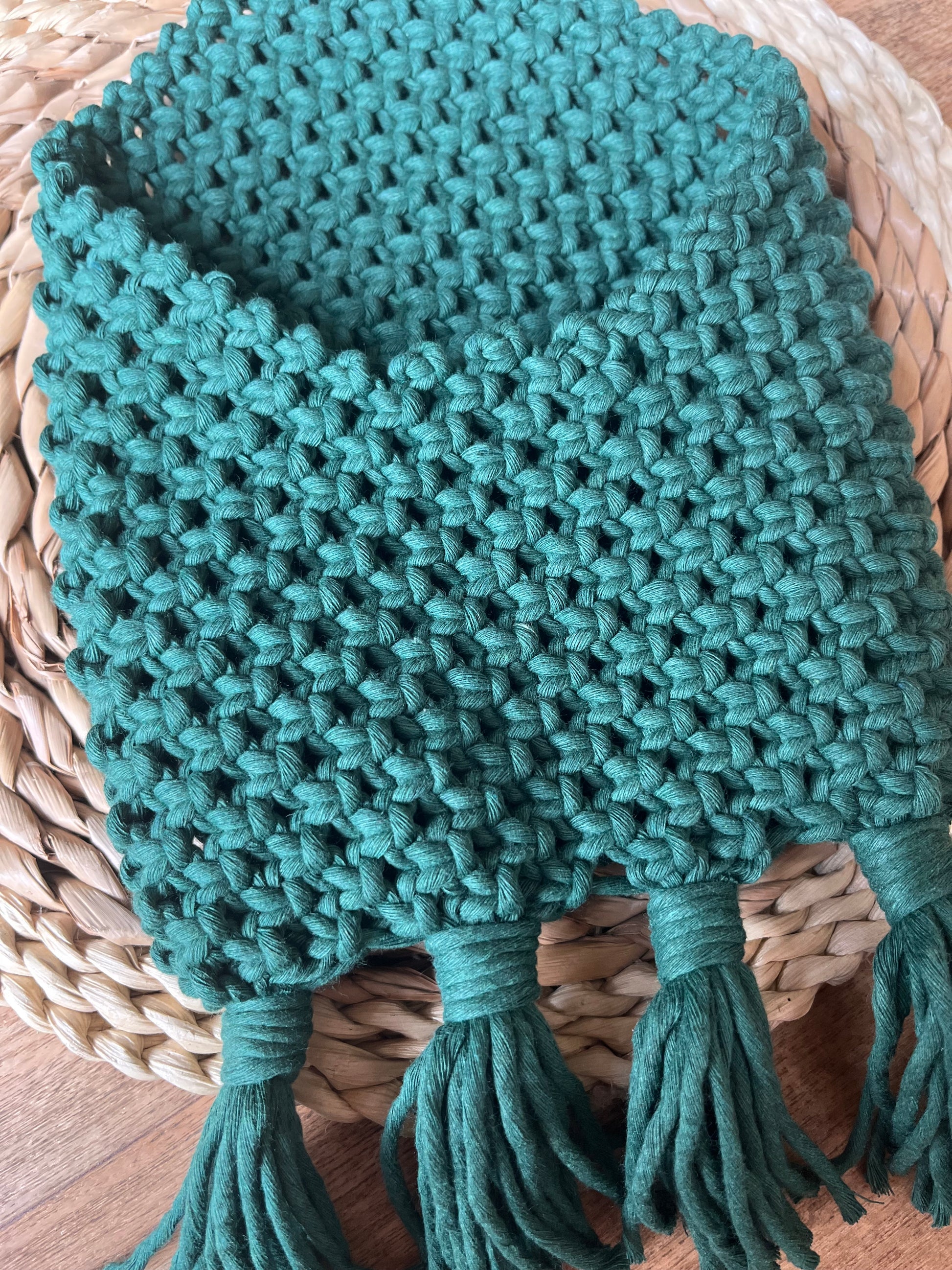 A hand-knitted, green scarf with a chunky texture is placed neatly on a woven straw surface, paired beautifully with a Macra-Made-With-Love Bedside storage pouch book sleeve. The scarf has several tassels hanging from the bottom edge, adding to its cozy and handmade appearance.