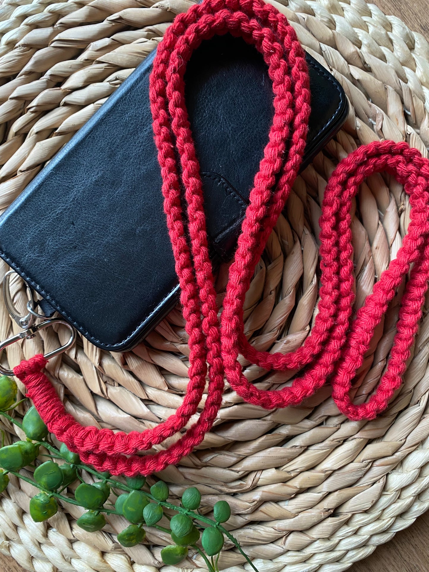 A red knitted lanyard, doubling as a handmade Macra-Made-With-Love Recycled Cotton Crossbody Phone Strap, is looped over a black wallet resting on a woven circular placemat. A small sprig of green plants lies near the bottom left corner of the wallet, all set against a slightly visible wooden surface.