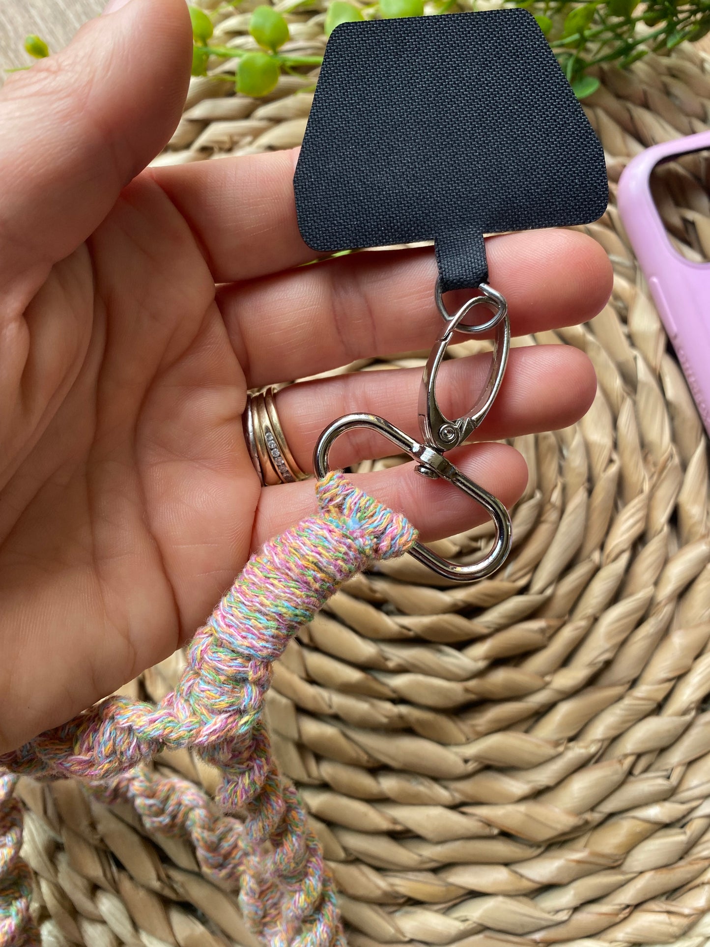 A hand holds a Macra-Made-With-Love Recycled cotton crossbody phone strap with a metal clip attached to a black plastic square. The colorful, eco-friendly lanyard rests on a woven basket surface, with greenery and a pink object visible in the background. The person is wearing a gold ring.