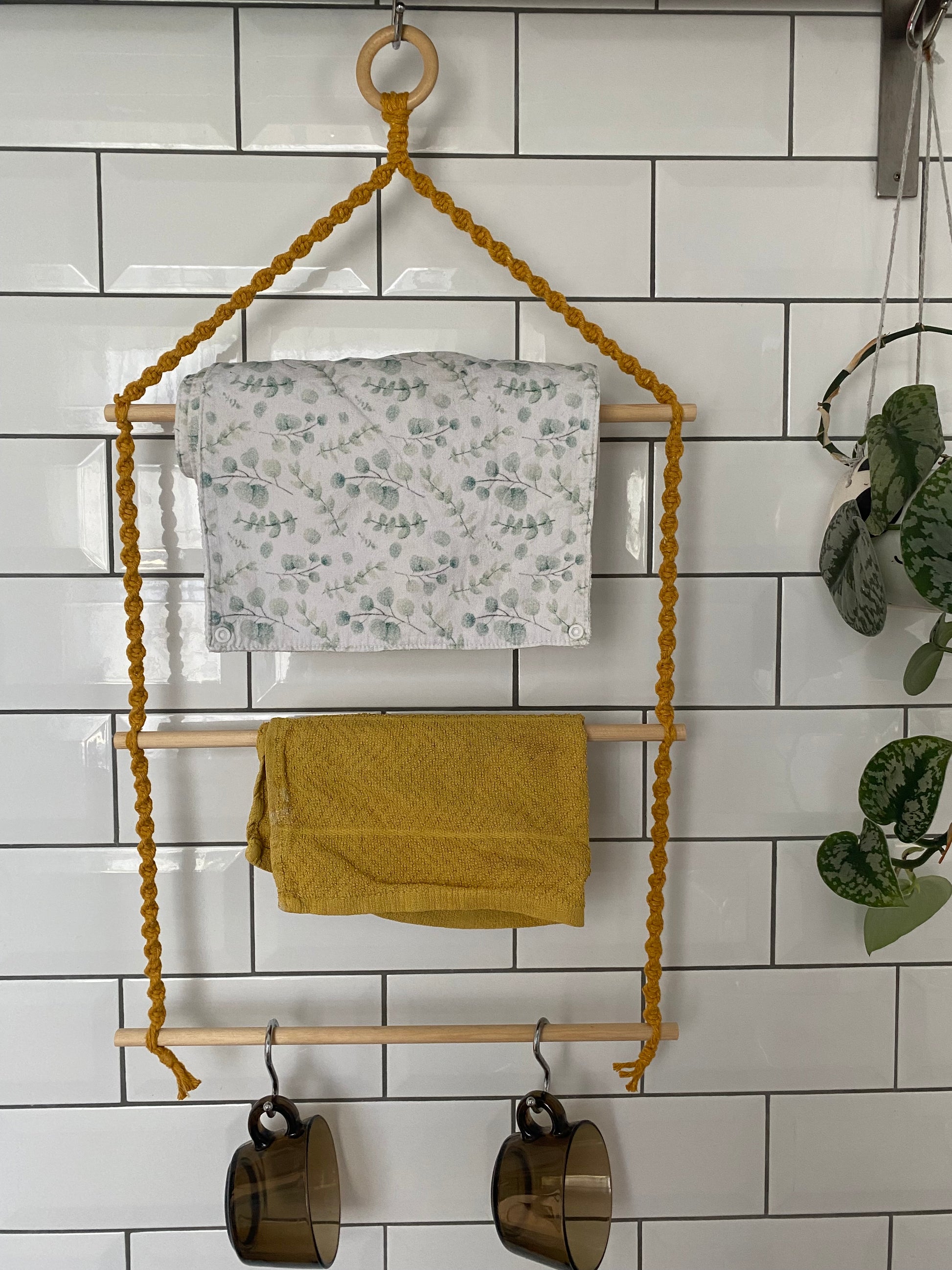A 3 tier hanging kitchen organiser made with a mustard yellow macrame rope, wooden ring and three wooden dowels hangs against a white tiled wall holding a white and green patterned cloth, a yellow tea towel and two brown mugs on each dowel.
