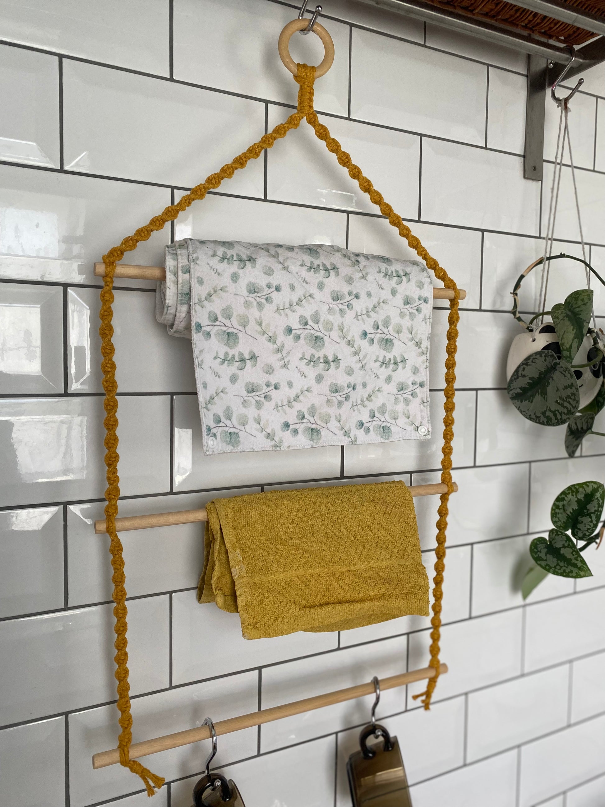 A handmade 3 tier kitchen organiser made with mustard yellow yarn, a wooden ring and three wooden dowels, hangs against a white tiled wall, holding one white tea towel with a green leaf pattern, one yellow tea towel and two glass brown mugs.