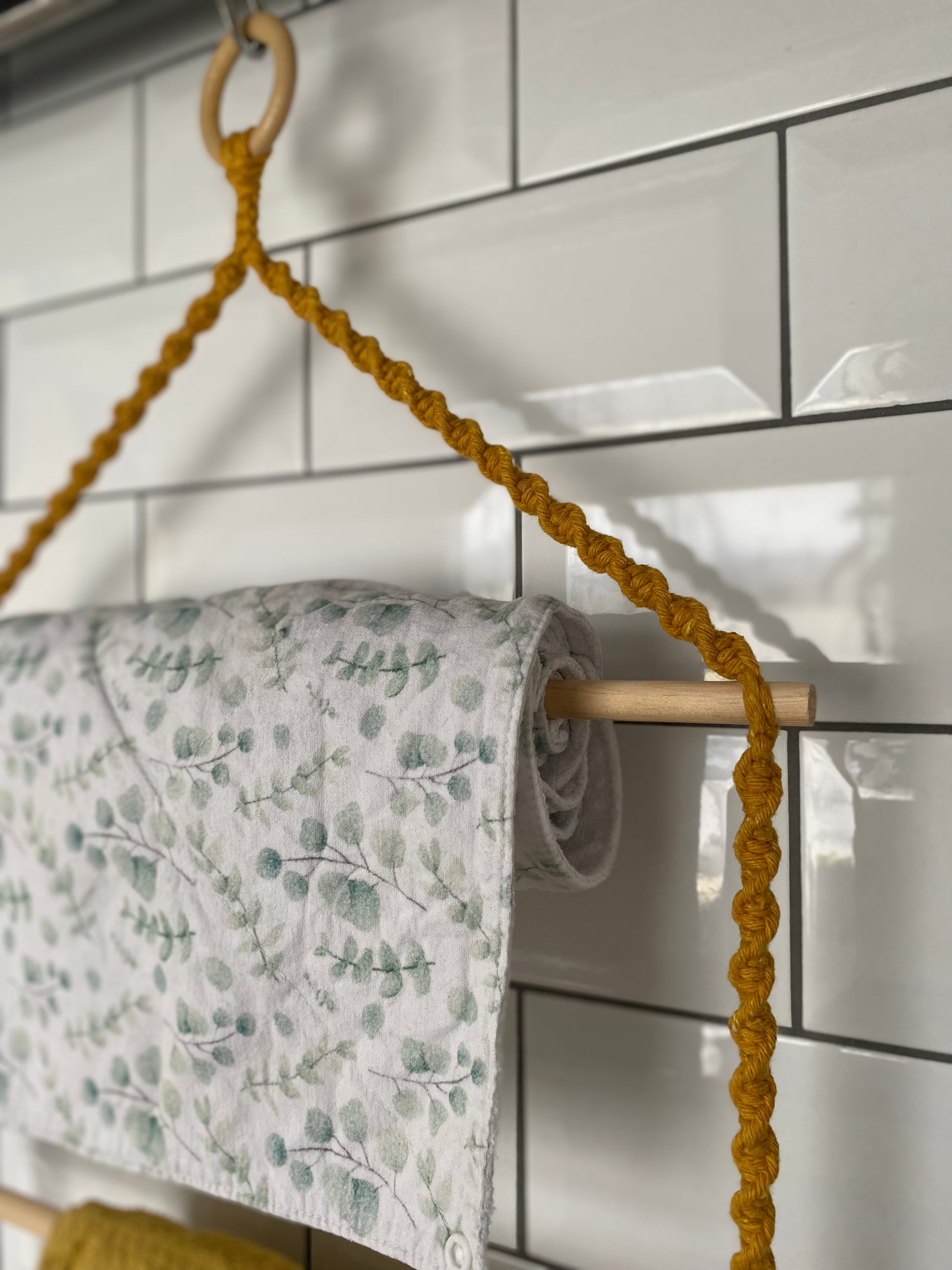 A close-up of a hanging kitchen organiser featuring a twisted mustard yellow macrame rope against a white tiled wall. A roll of white and green fabric towel is draped on the top dowel of the hanger.