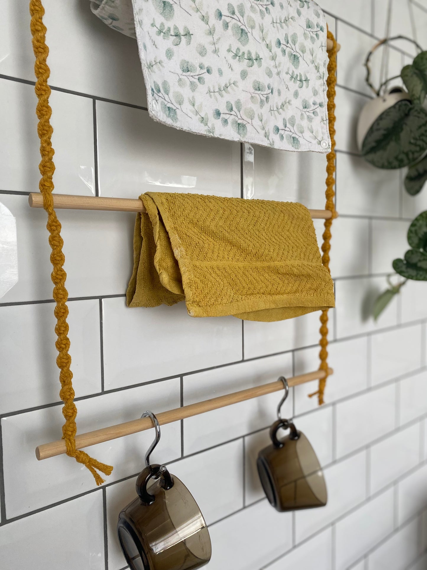 A hanging kitchen organiser with mustard yellow macrame ropes and wooden dowels, hangs against a white tiled wall. The wooden organiser holds two cloths, one with a green leafy pattern and one in mustard yellow, and two brown glass mugs hang on separate wooden dowels.