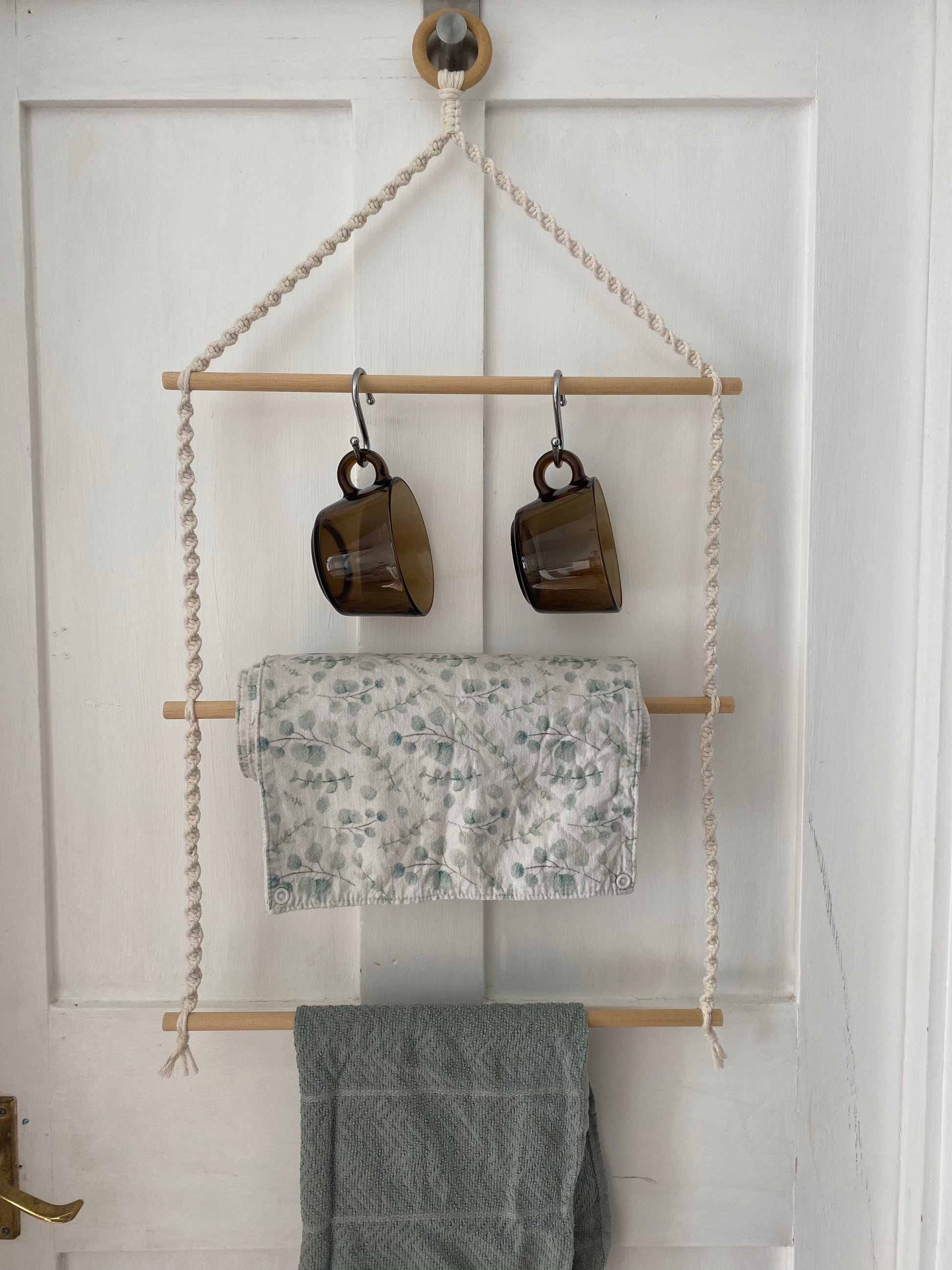A 3 tier wooden kitchen organiser mounted on a white door, from a wooden ring. The top dowel holds two brown glass mugs, the middle shelf has a roll of floral-patterned cloth and a green cotton tea towel hangs below on the third wooden dowel.