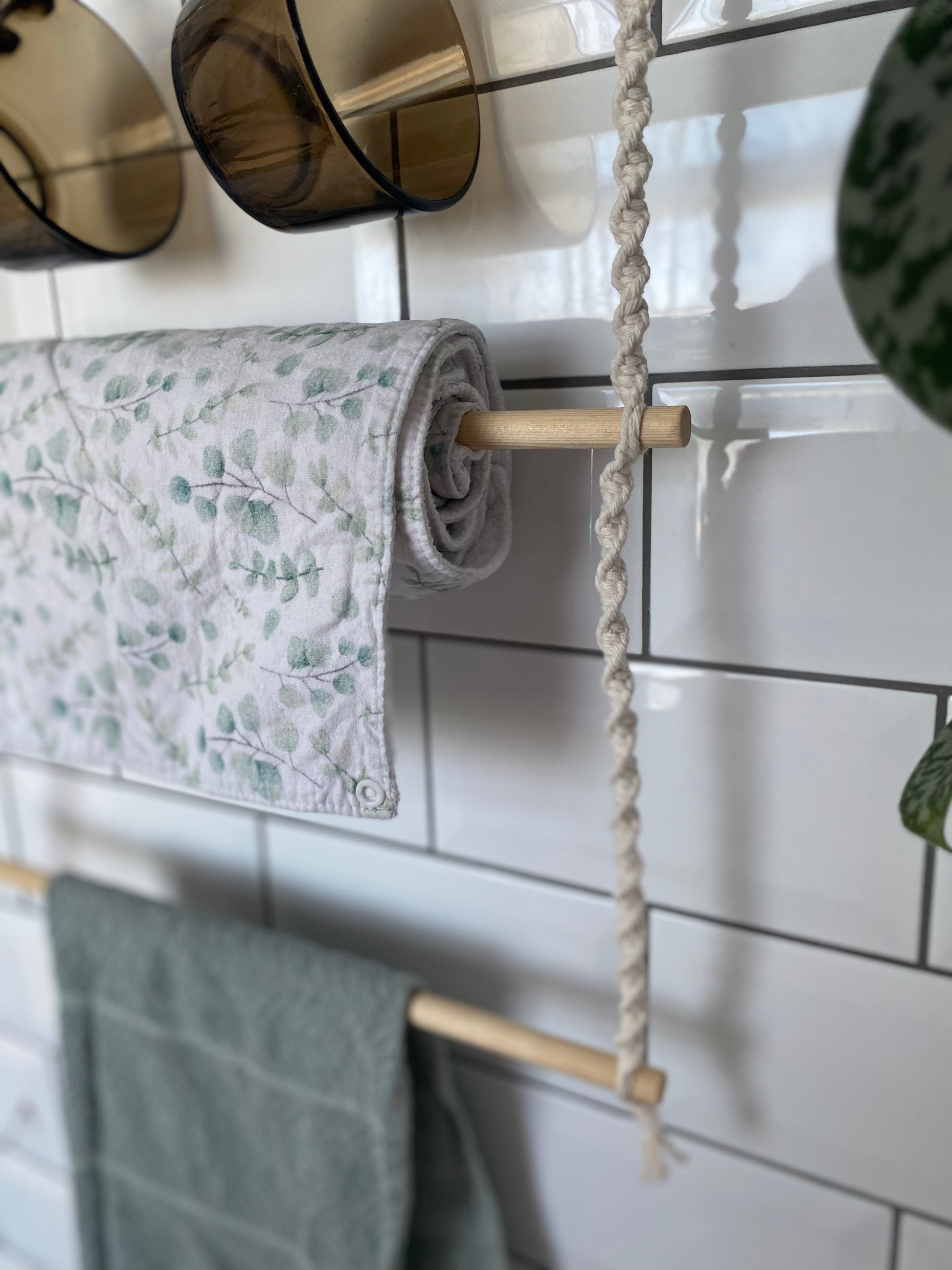 A close-up of a hanging organiser made with natural cotton and wooden rods, holding a roll of white and green patterned fabric towel, a green towel and two brown glass mugs against a white tiled wall.