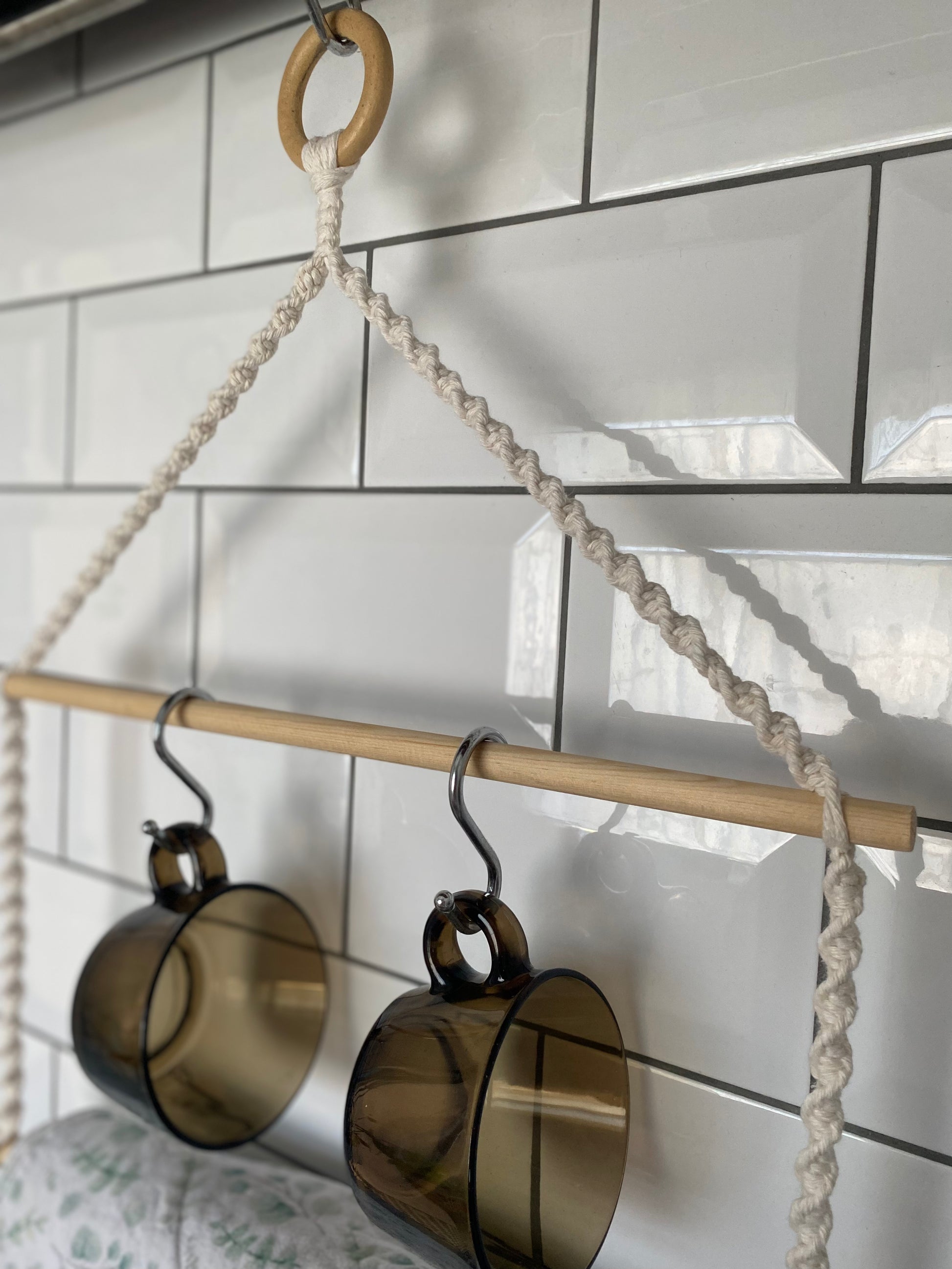 Close-up of two amber-coloured glass mugs hanging by their handles on metal S hooks from a wooden rod, which is suspended by a twisted rope made of recycled cotton with a wooden ring at the top.