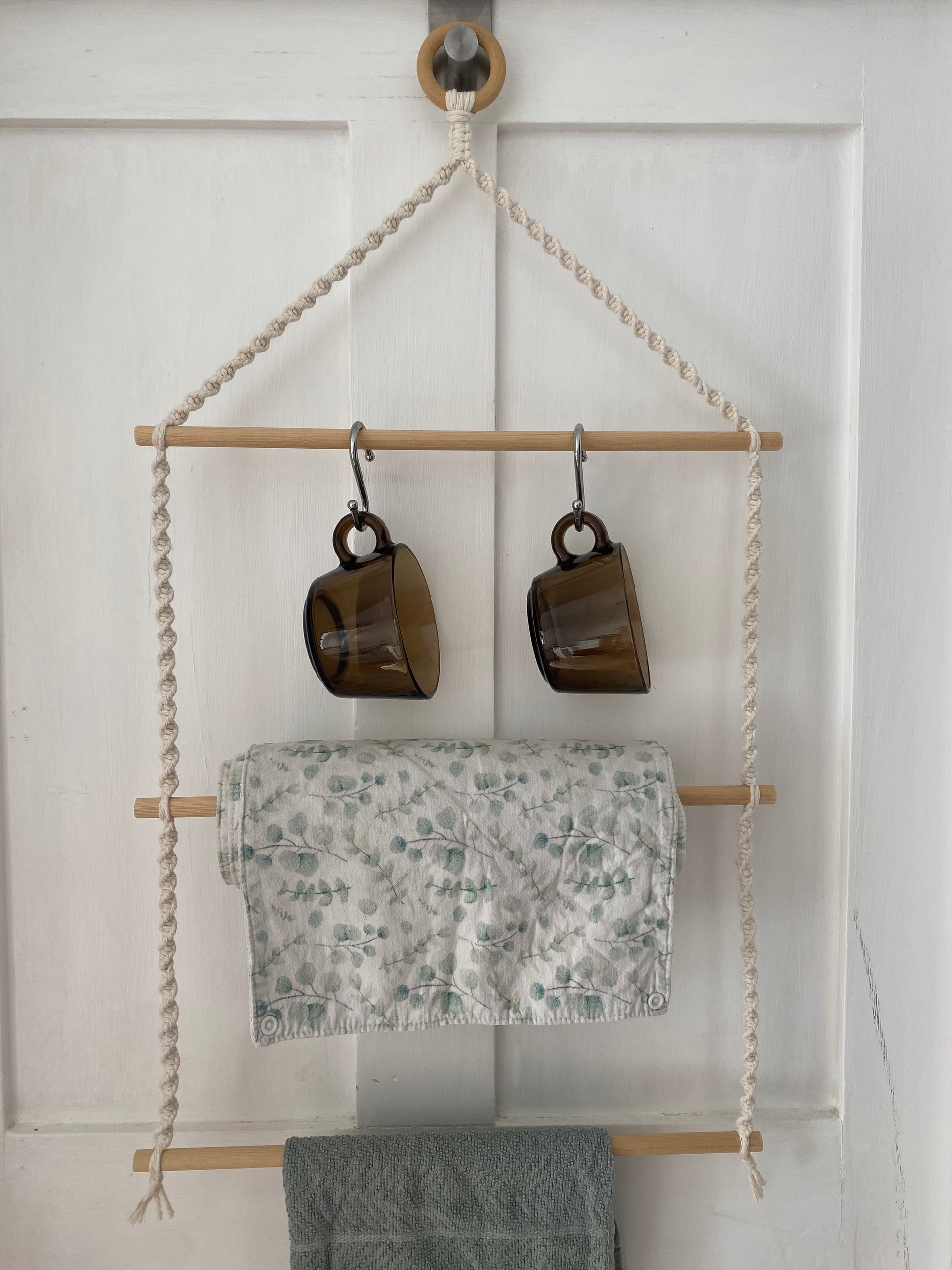 A handmade 3 tier wooden kitchen organiser, made with natural cotton and wooden dowels. Two brown glass cups on metal S hooks are hanging on the top dowel, a folded leafy-patterned cloth in the middle, and a green tea towel is draped below on the bottom dowel.
