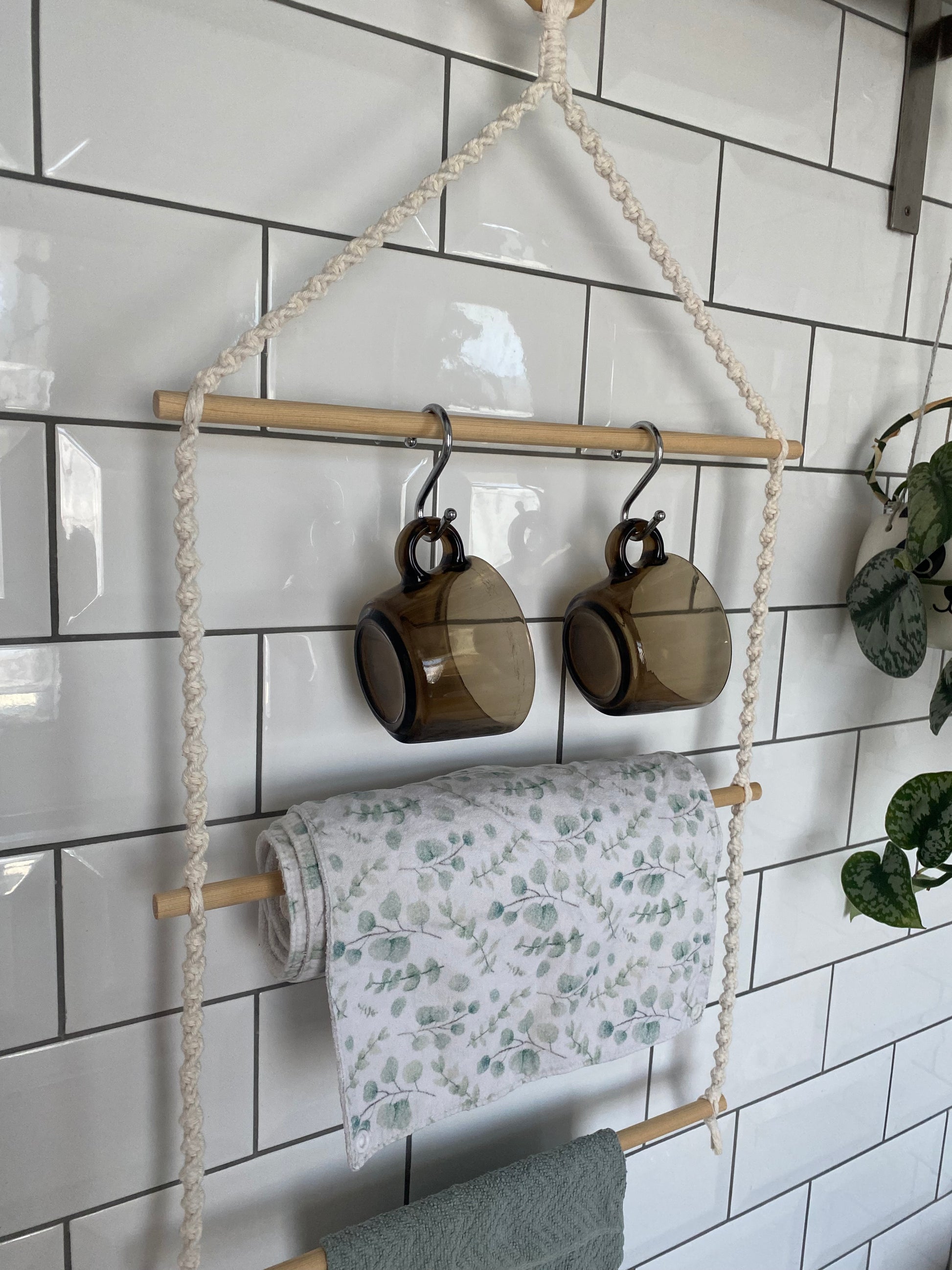 A 3 tier kitchen organiser with natural rope and wooden dowels hangs against a white tied wall. Two brown glass mugs hang on metal S hooks from the top rod, a white and green leaf-patterned cloth is draped over the middle rod, and a green towel is just seen on the bottom rod.