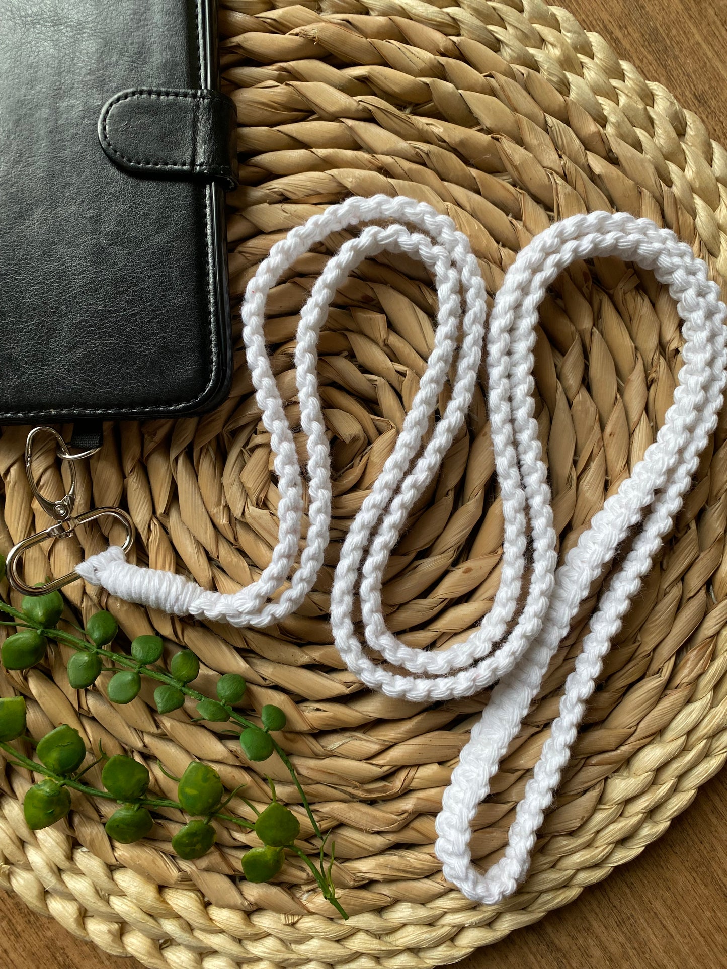 An eco-friendly, braided white lanyard lies coiled on a woven placemat. Next to it is a black leather-bound notebook with a clasp, partially visible. A small green plant accent is placed near the bottom left corner of the image, complementing the Macra-Made-With-Love Recycled Cotton Crossbody Phone Strap beautifully.