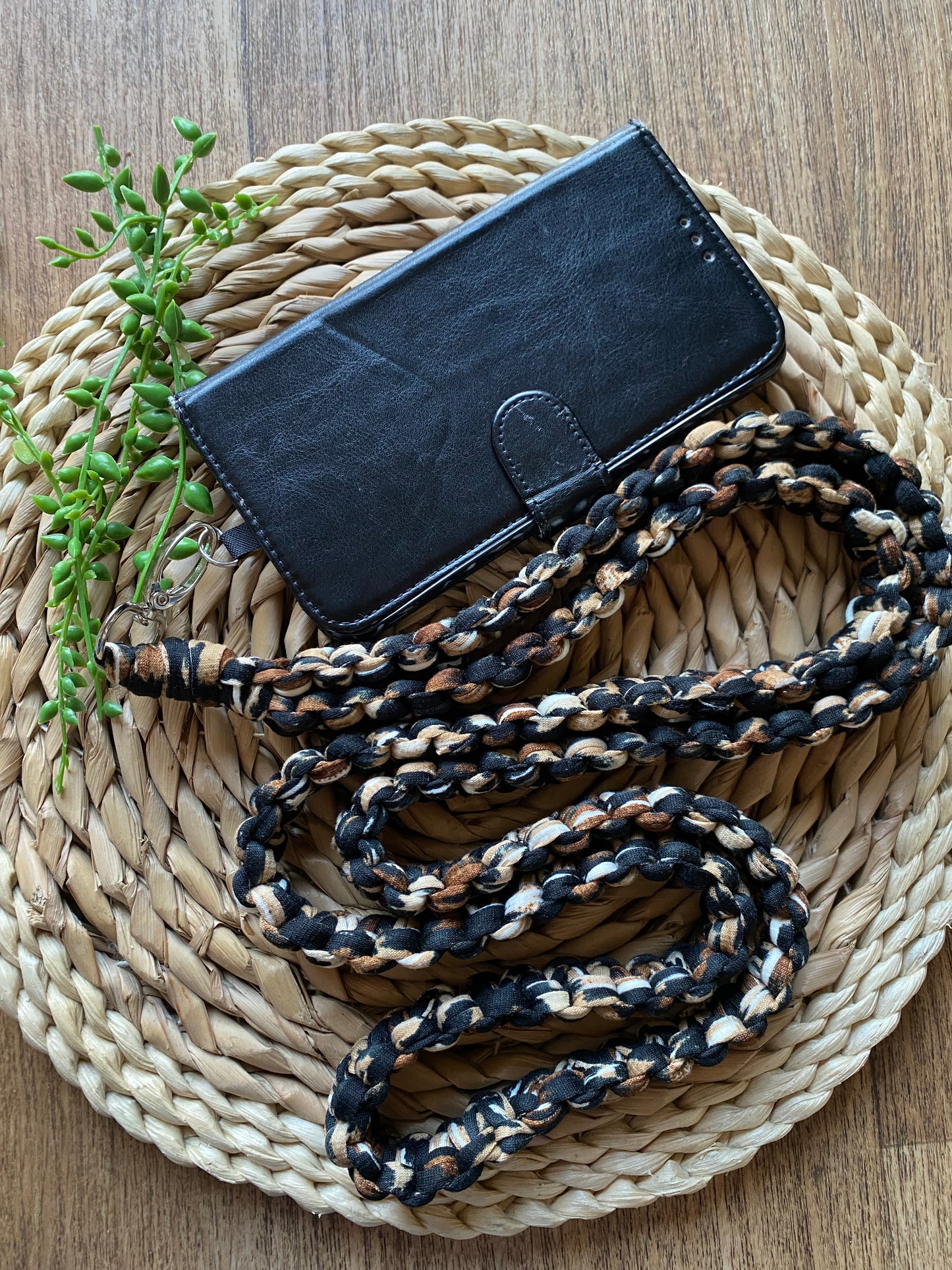 A black leather phone wallet with a snap closure rests on a round wicker mat. Attached to the wallet is an eco-friendly, handmade Macra-Made-With-Love Recycled cotton crossbody phone strap in black, brown, and beige colors. A small green plant decorates the upper left side of the mat.