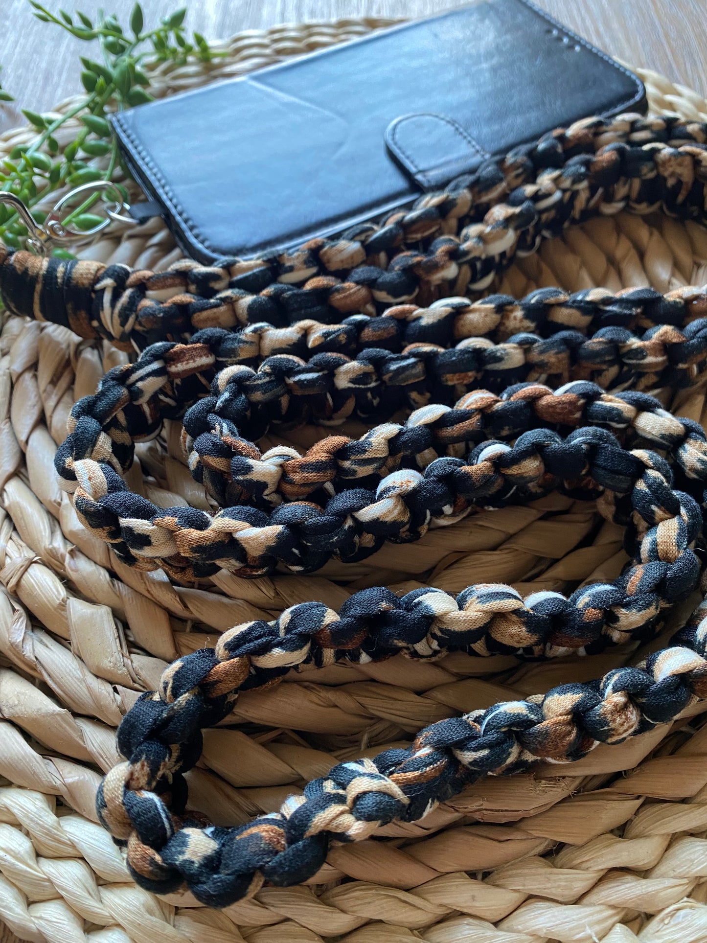 A braided, multicolored fabric rope—an eco-friendly, handmade Recycled cotton crossbody phone strap by Macra-Made-With-Love—is coiled on a straw-textured mat. Behind it is a black leather wallet and a small green plant, adding nature to the rugged yet elegant composition.