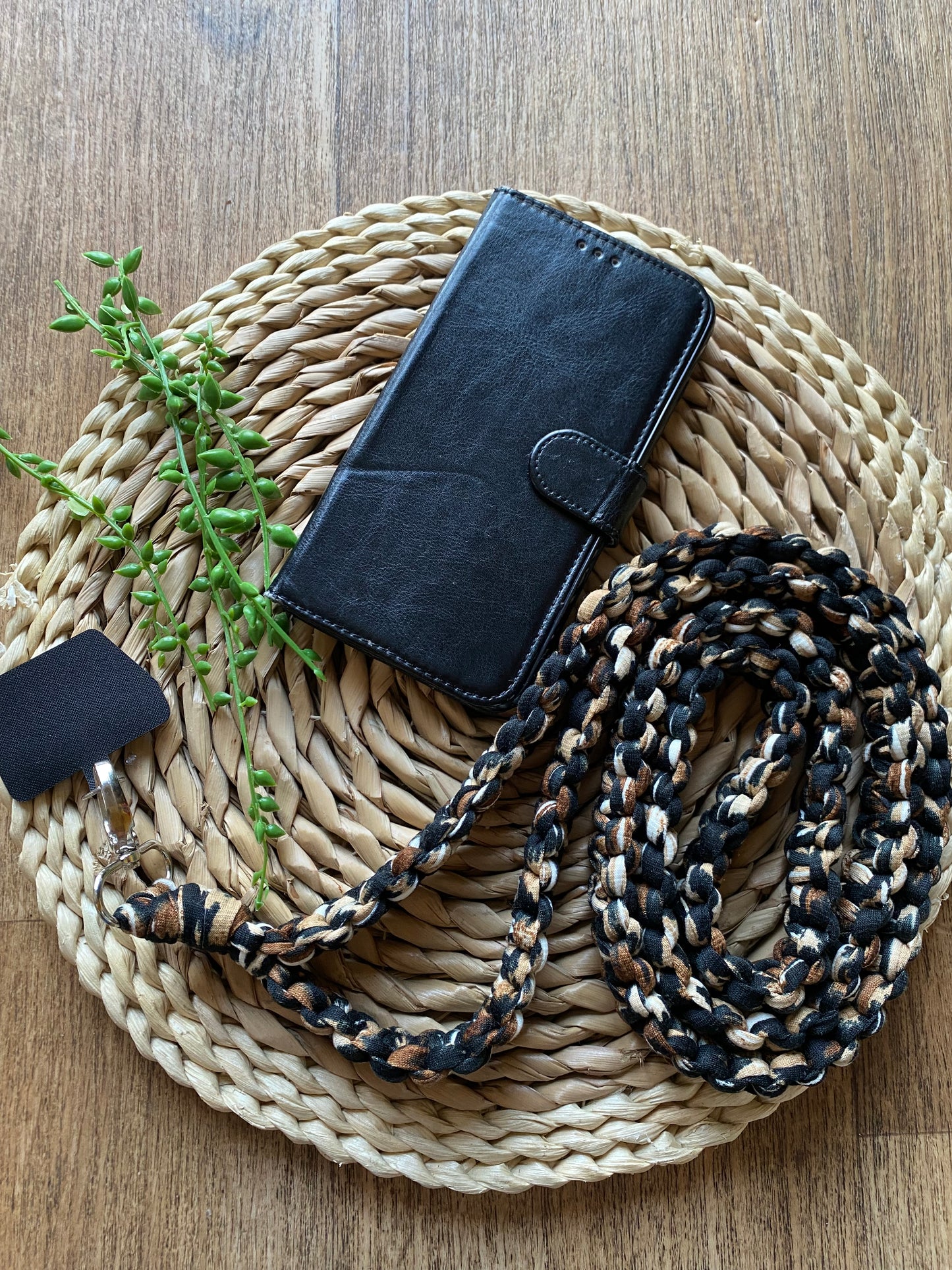 A black wallet-style phone case with a magnetic clasp lies on a woven placemat. Next to it is an eco-friendly, black and brown braided lanyard with a keychain attached, perfectly complementing the Macra-Made-With-Love Recycled cotton crossbody phone strap. A green leafy stem decorates the wooden surface background.