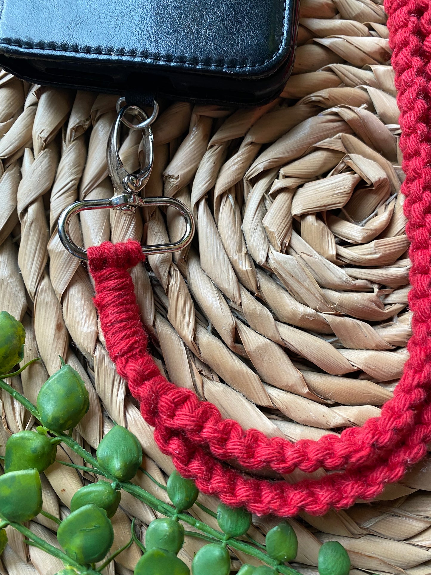 A black leather wallet with a metal clasp is attached to a red Recycled cotton crossbody phone strap by Macra-Made-With-Love on a textured, circular woven mat. Green artificial leaves are partially visible in the bottom left corner, adding a touch of eco-friendly charm.