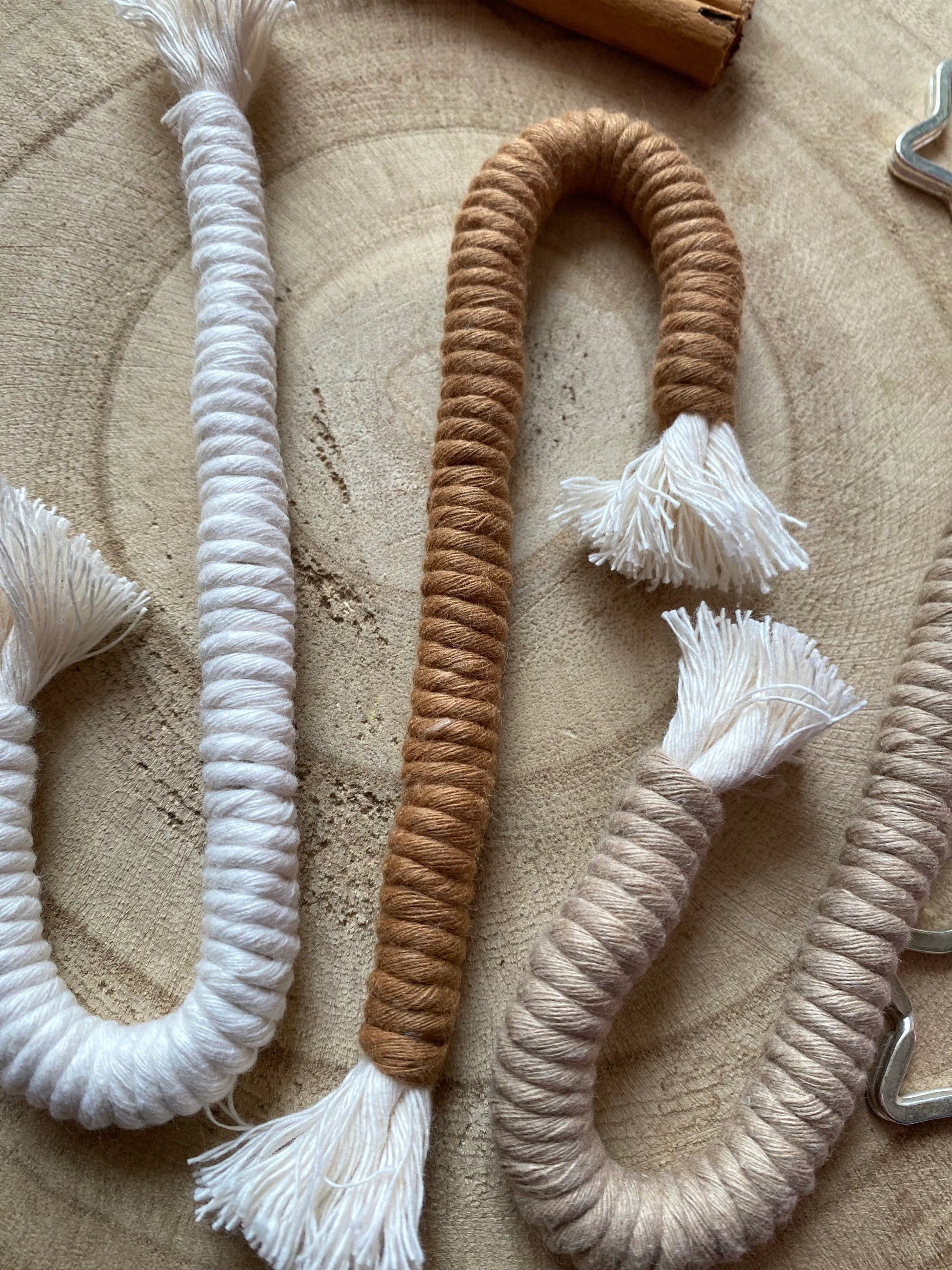 A close up image of a set of three Christmas candy cane shaped macrame ornaments in beige, white, and brown colours with tassels, displayed on a wooden surface.