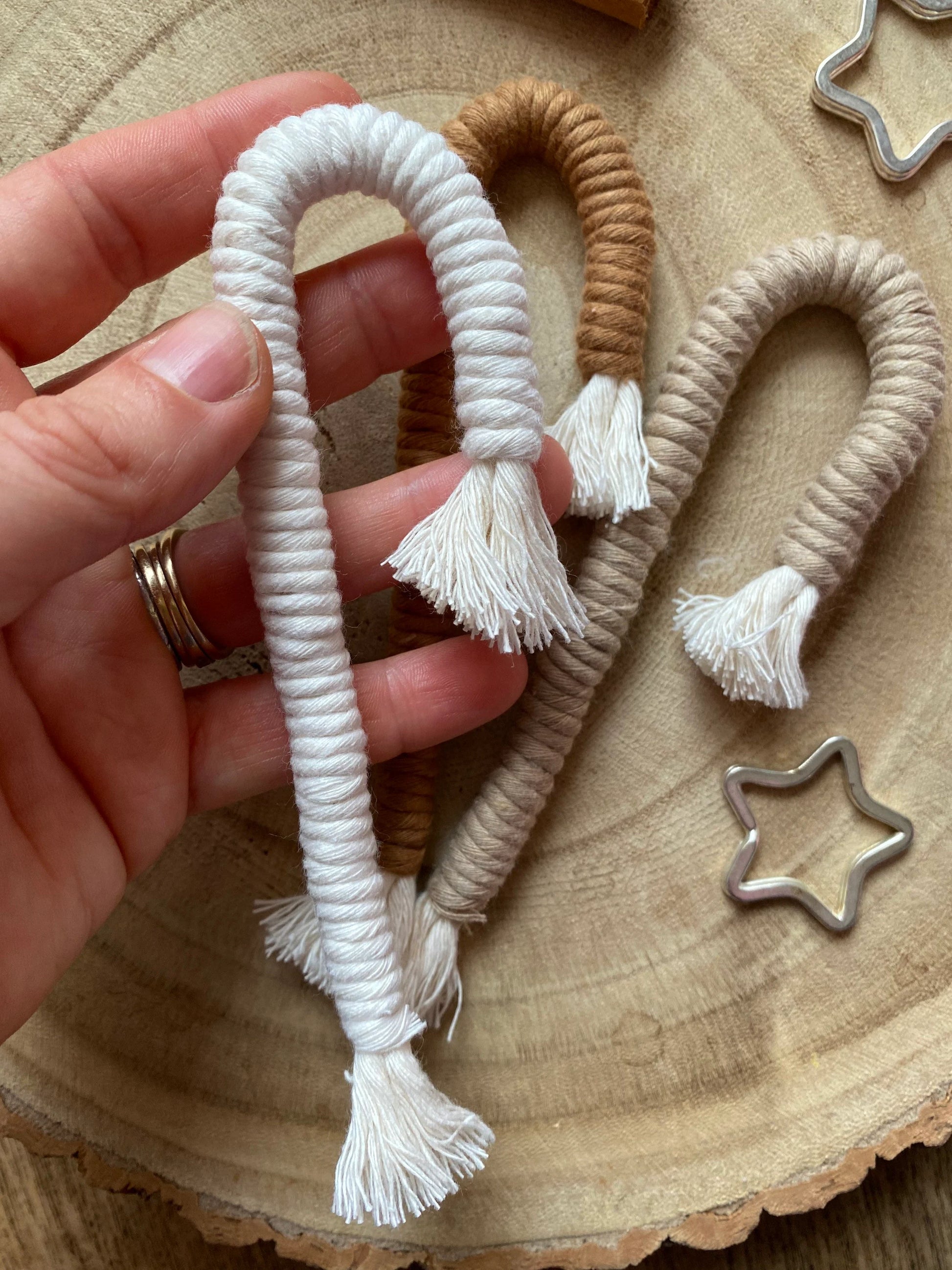 A hand holding a white candy cane shaped macrame ornament, there are two further candy cane shaped ornaments on a wooden surface below, in beige and brown colours, each with white tassels.