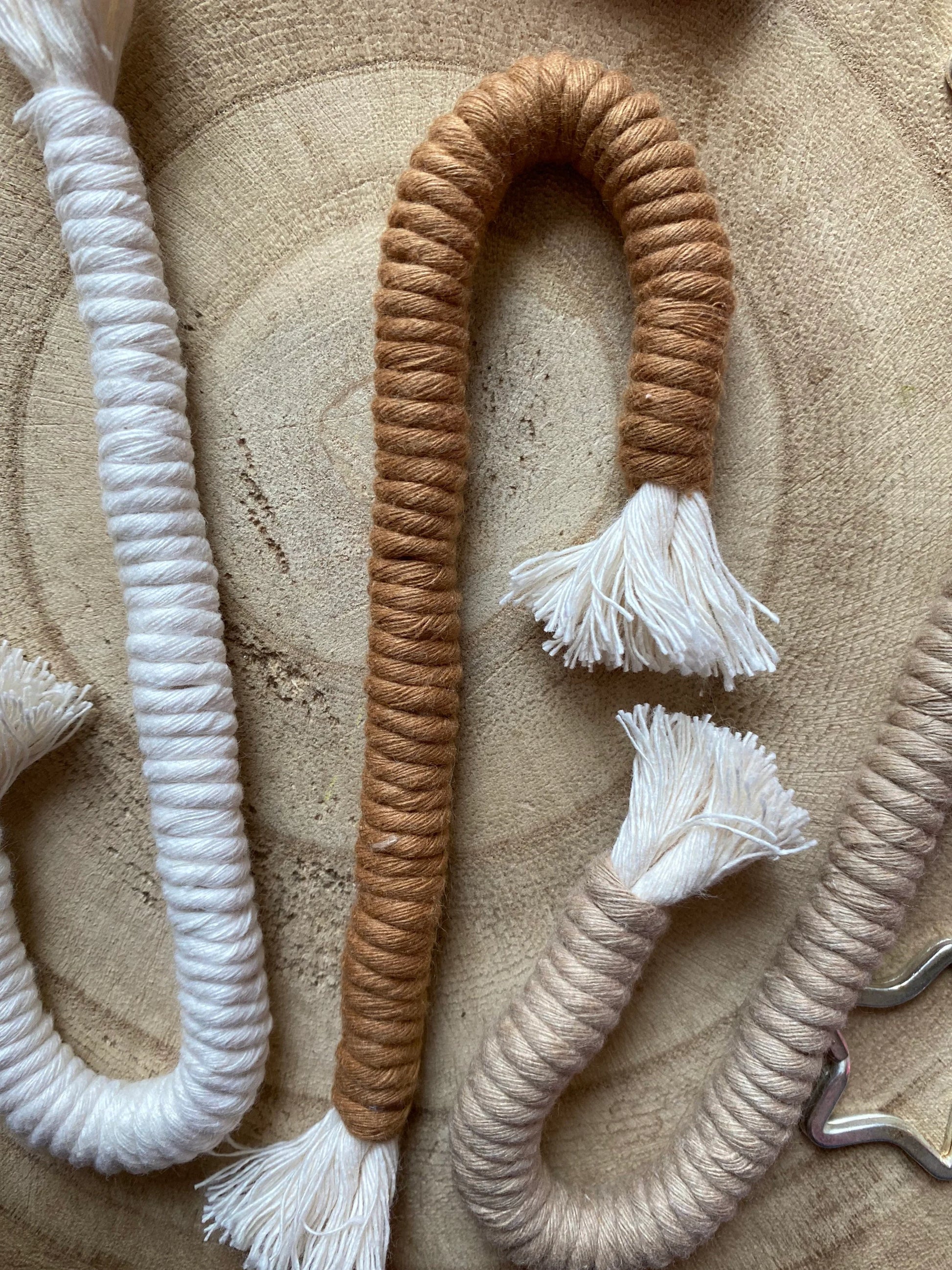 A close up image of a set of three Christmas candy cane shaped macrame ornaments in beige, white, and brown colours with tassels, displayed on a wooden surface.