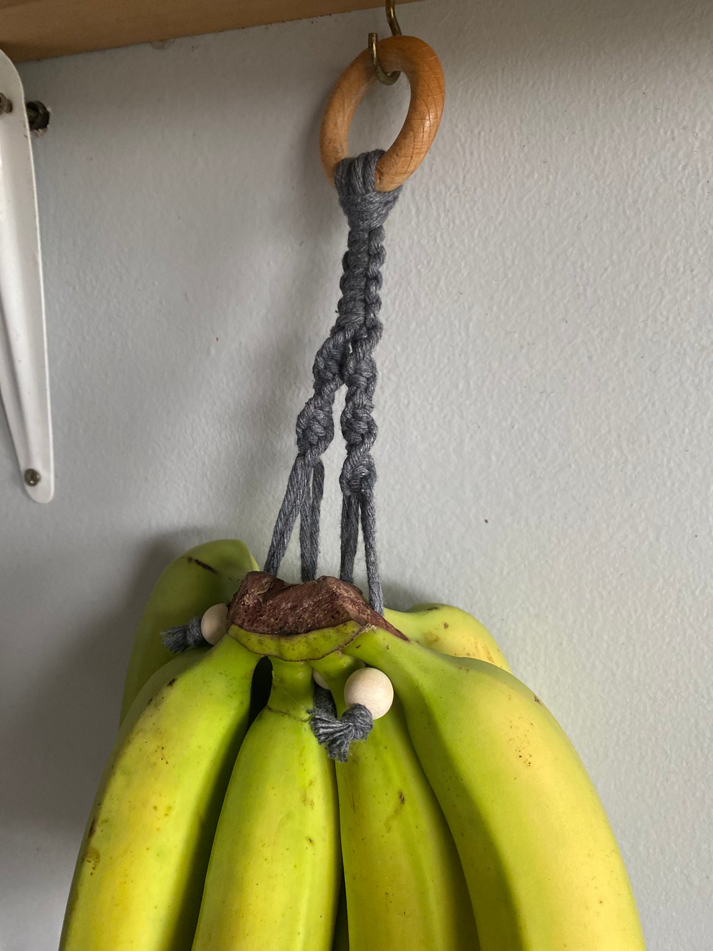 A bunch of green bananas hangs from a Macra-Made-With-Love Banana Hanger with a wooden ring at the top. The hanger, exemplifying eco-friendly home decor, is attached to a metal hook on the underside of a wooden shelf. The bananas are still unripe with firm, green skin.