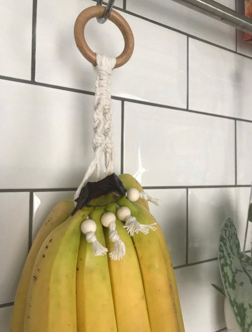 A bunch of ripe bananas hangs from a Macra-Made-With-Love Banana Hanger with white beads, attached to a wooden ring on a tiled wall. This handmade kitchen decor adds a decorative touch while keeping the bananas elevated.