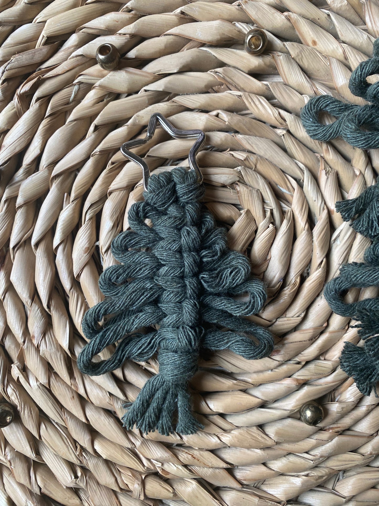 A small, dark green Knotted Yarn Hanging Christmas Tree Decoration from Macra-Made-With-Love lies on a woven straw mat. Crafted from recycled cotton yarns, the tree features intricate knotted loops and is topped with a metal star, making it an eco-friendly holiday decor piece.