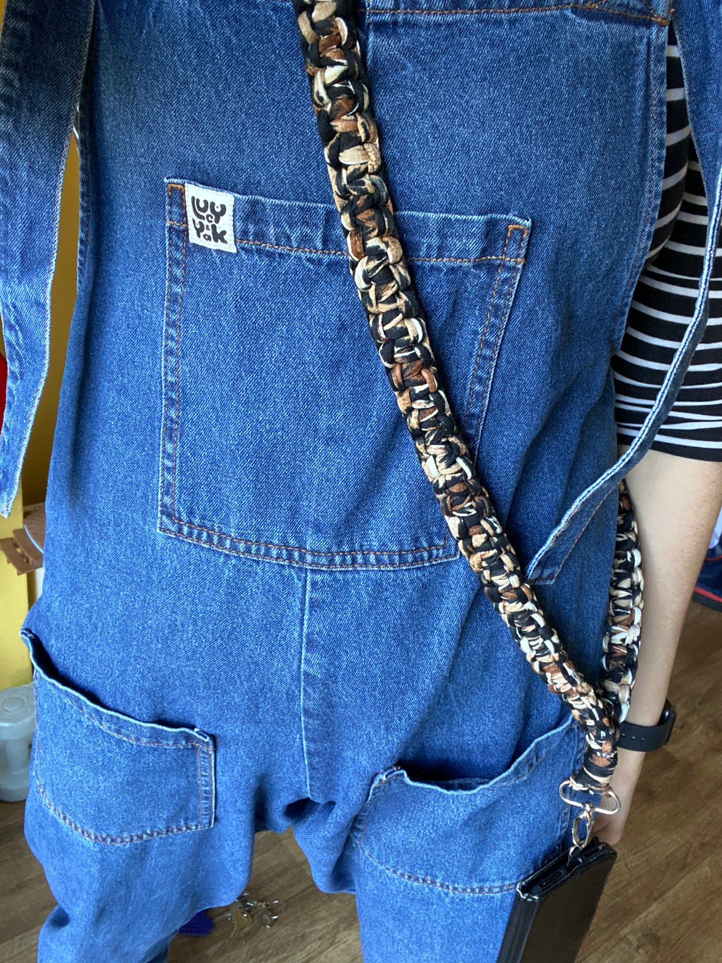 Person wearing blue denim overalls and a black and white striped shirt underneath. They have a braided, eco-friendly handmade Recycled cotton crossbody phone strap by Macra-Made-With-Love over their shoulder and an enamel pin on the front pocket of the overalls.