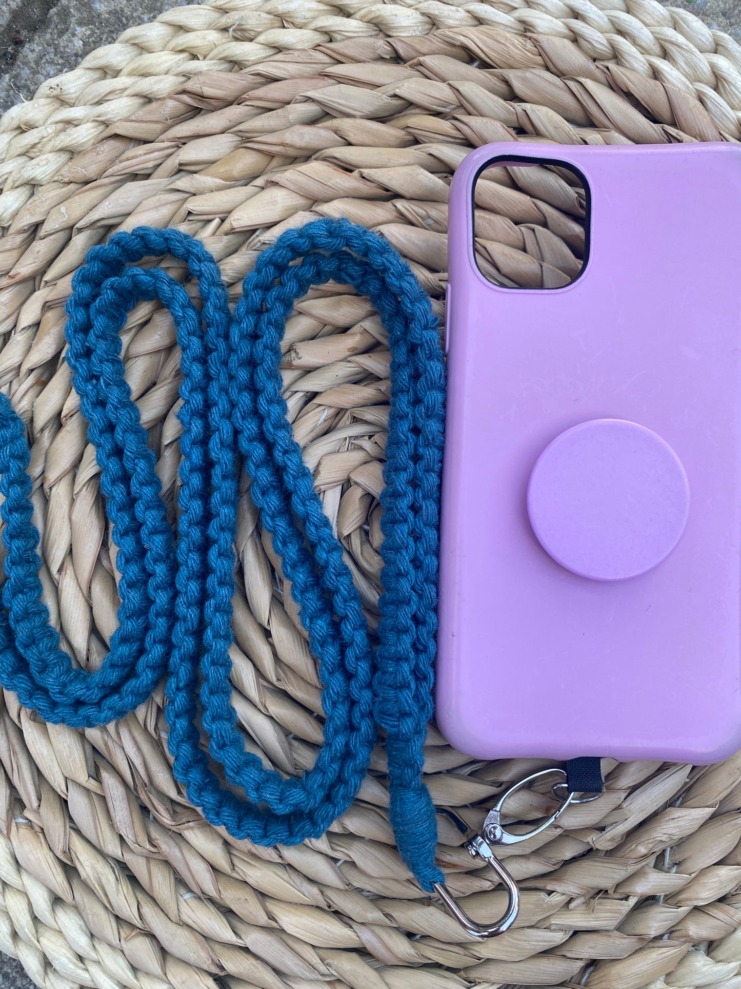 A coiled blue knitted strap with a metal clip, possibly for holding or securing an item, is placed next to a purple phone case with a circular pop socket on a woven straw mat. The setup radiates charm with the eco-friendly **Macra-Made-With-Love Recycled cotton crossbody phone strap** as the standout phone accessory.