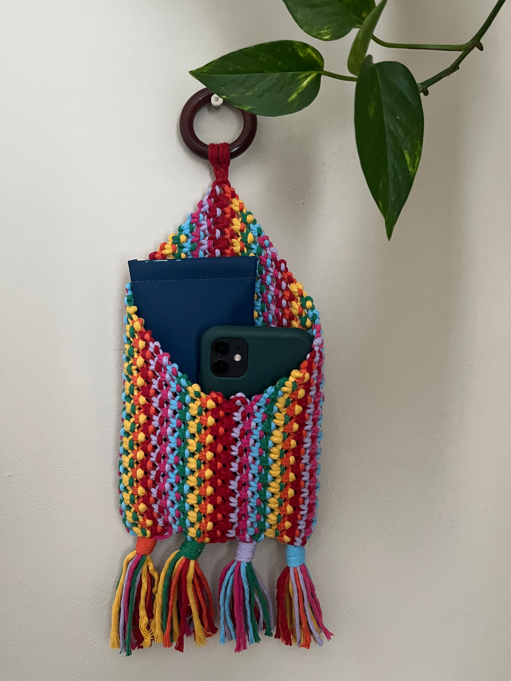 A colorful, handmade Macra-Made-With-Love Bedside Storage Pouch Book Sleeve with a wooden ring at the top is hanging on a white wall. The pouch, made from recycled cotton decor, holds a blue notebook and a green smartphone. It features bright stripes and tassels at the bottom. A leafy plant is visible on the right.