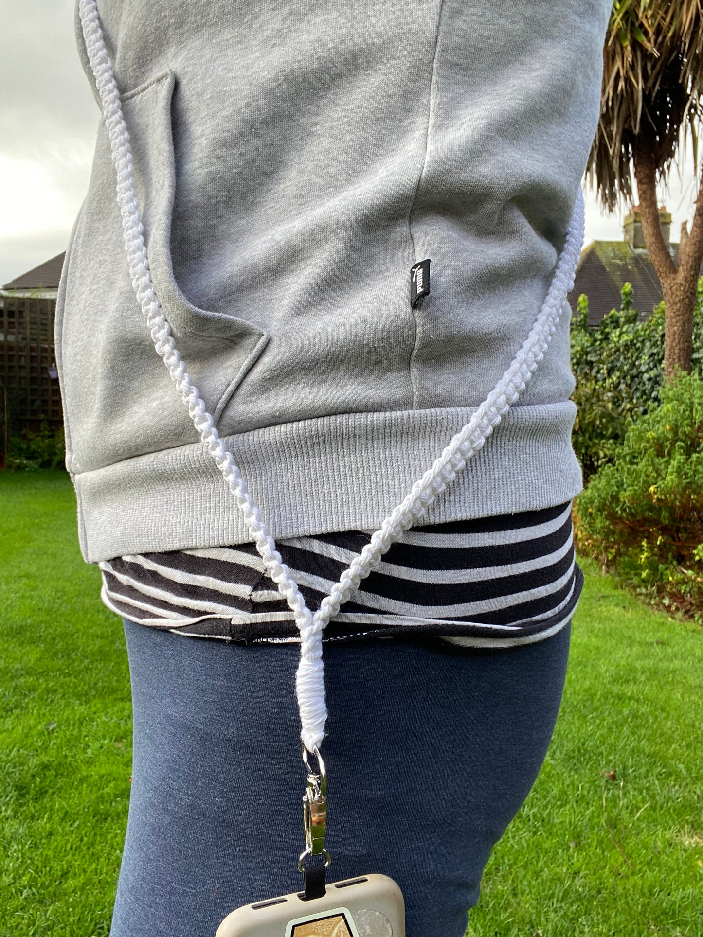 Person wearing a grey hoodie and blue pants, standing outdoors on a grassy lawn with trees and shrubs in the background. A phone is attached to a white cord connected to the hoodie, featuring an eco-friendly handmade Macra-Made-With-Love Recycled Cotton Crossbody Phone Strap.