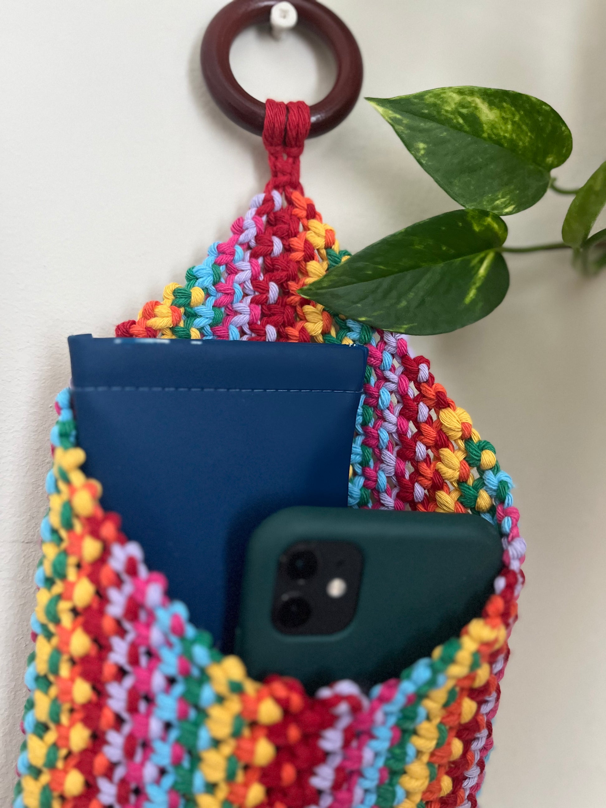 A colorful crocheted wall pouch made from recycled cotton hangs from a circular hook. The eco-friendly Macra-Made-With-Love Bedside storage pouch book sleeve contains a blue notebook and a smartphone with a dark green case. A green leafy plant is visible in the background, adding a touch of nature to the scene.