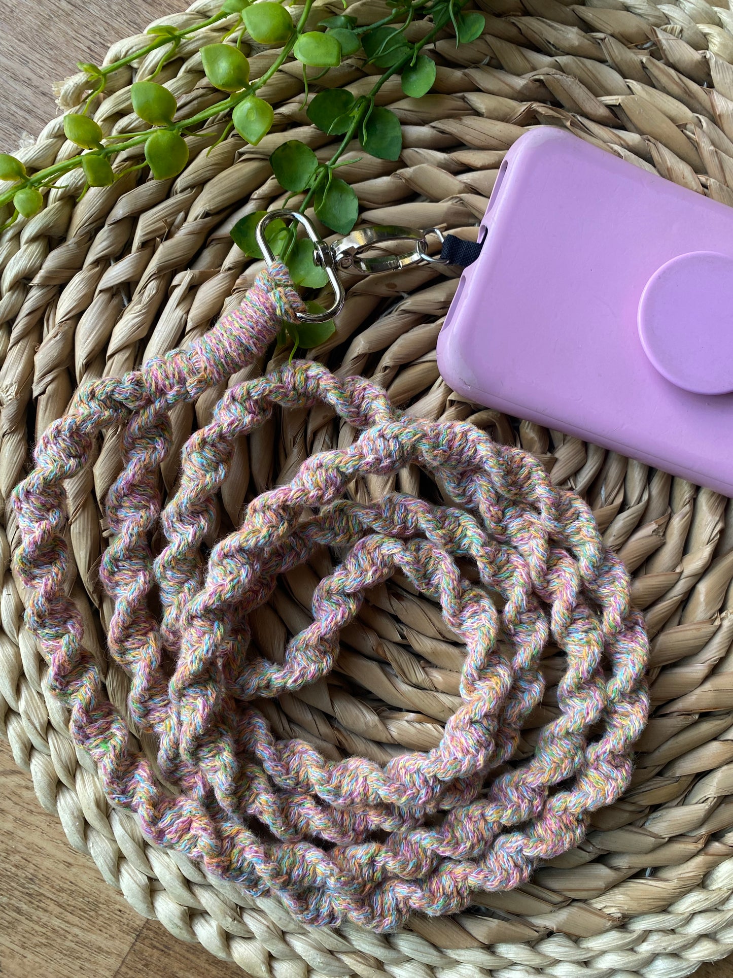 A braided, multicolored phone accessory with a silver clip is attached to a pink phone case. The **Macra-Made-With-Love Recycled cotton crossbody phone strap** and phone case rest on a woven, round placemat, with some small green plant leaves in the background.