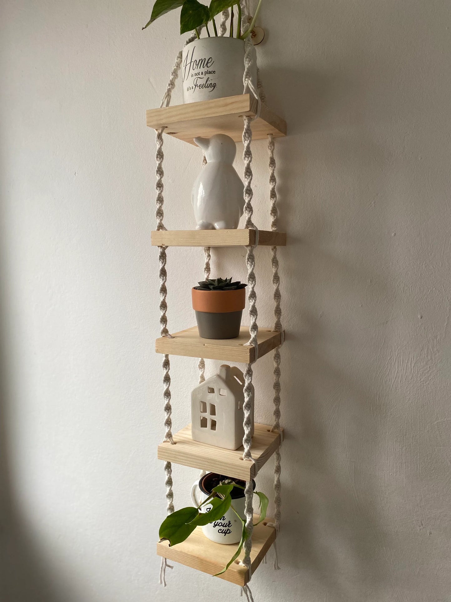 Handmade macrame rope shelves, made with small square wooden shelves, held together with natural knotted rope. Displayed hanging on a white wall, with small houseplants on ornaments displayed on each shelf.