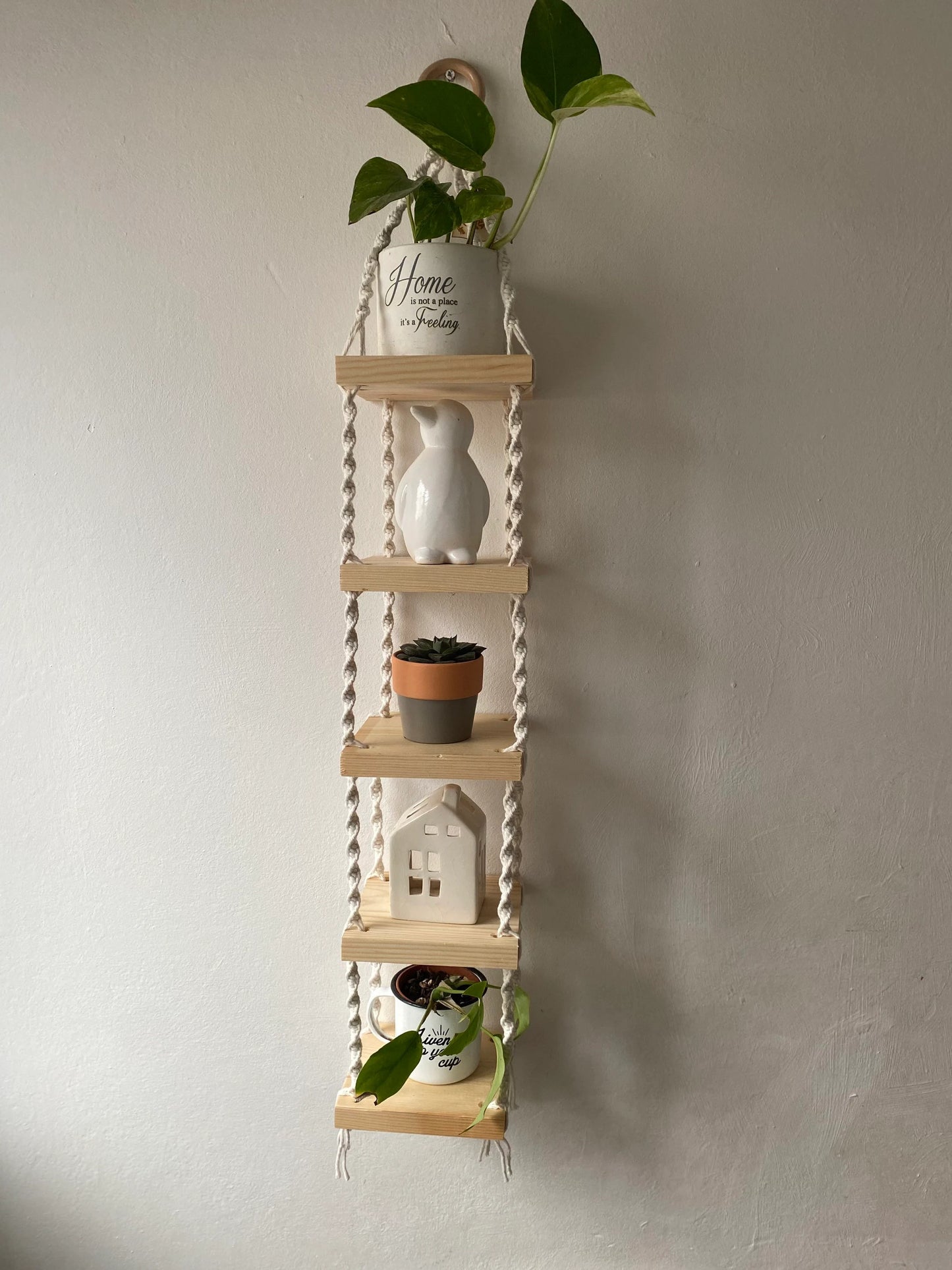 Handmade macrame rope shelves, made with small square wooden shelves, held together with natural knotted rope. Displayed hanging on a white wall, with small houseplants on ornaments displayed on it.
