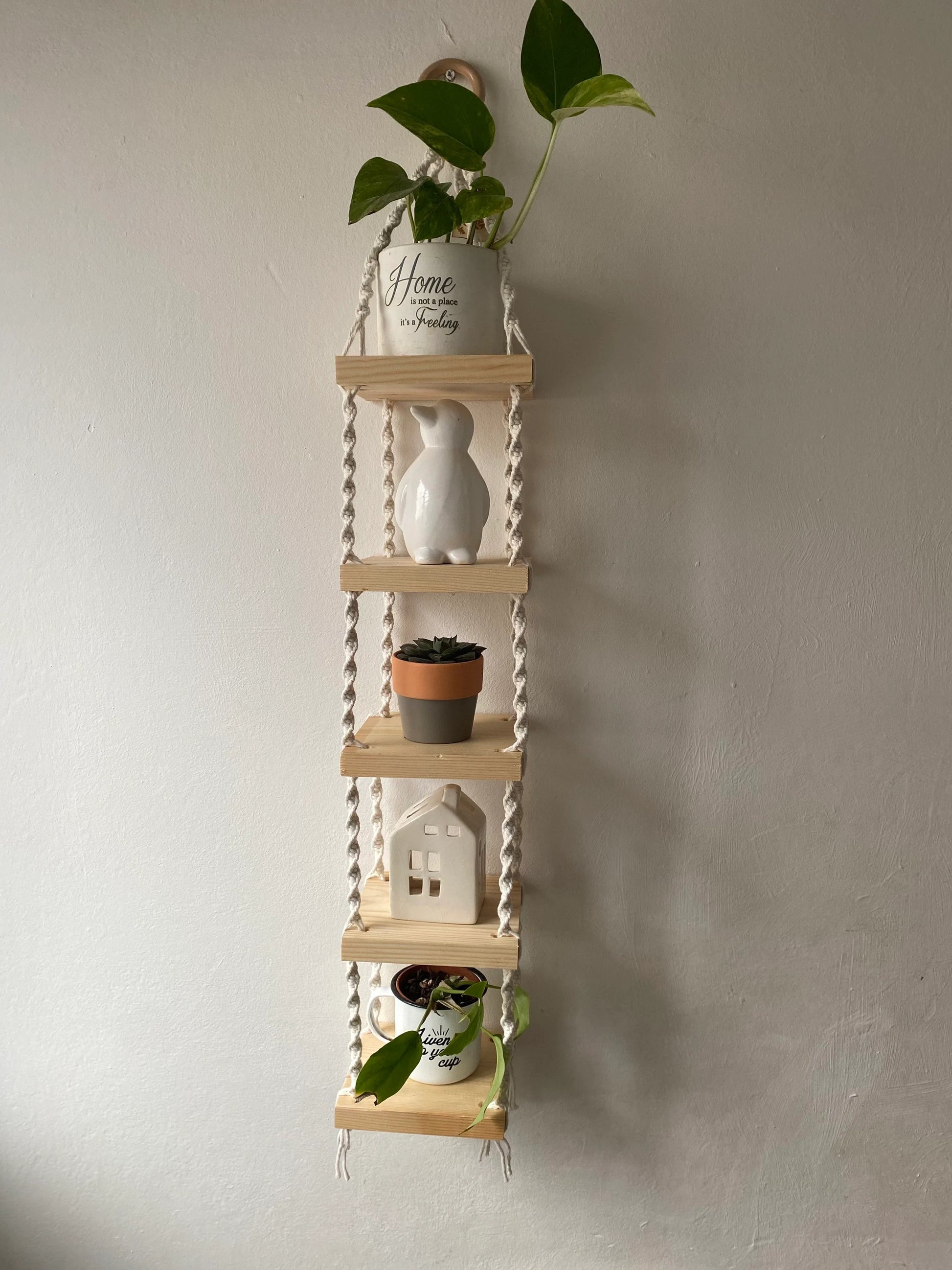Handmade macrame rope shelves, made with small square wooden shelves, held together with natural knotted rope. Displayed hanging on a white wall, with small houseplants on ornaments displayed on it.