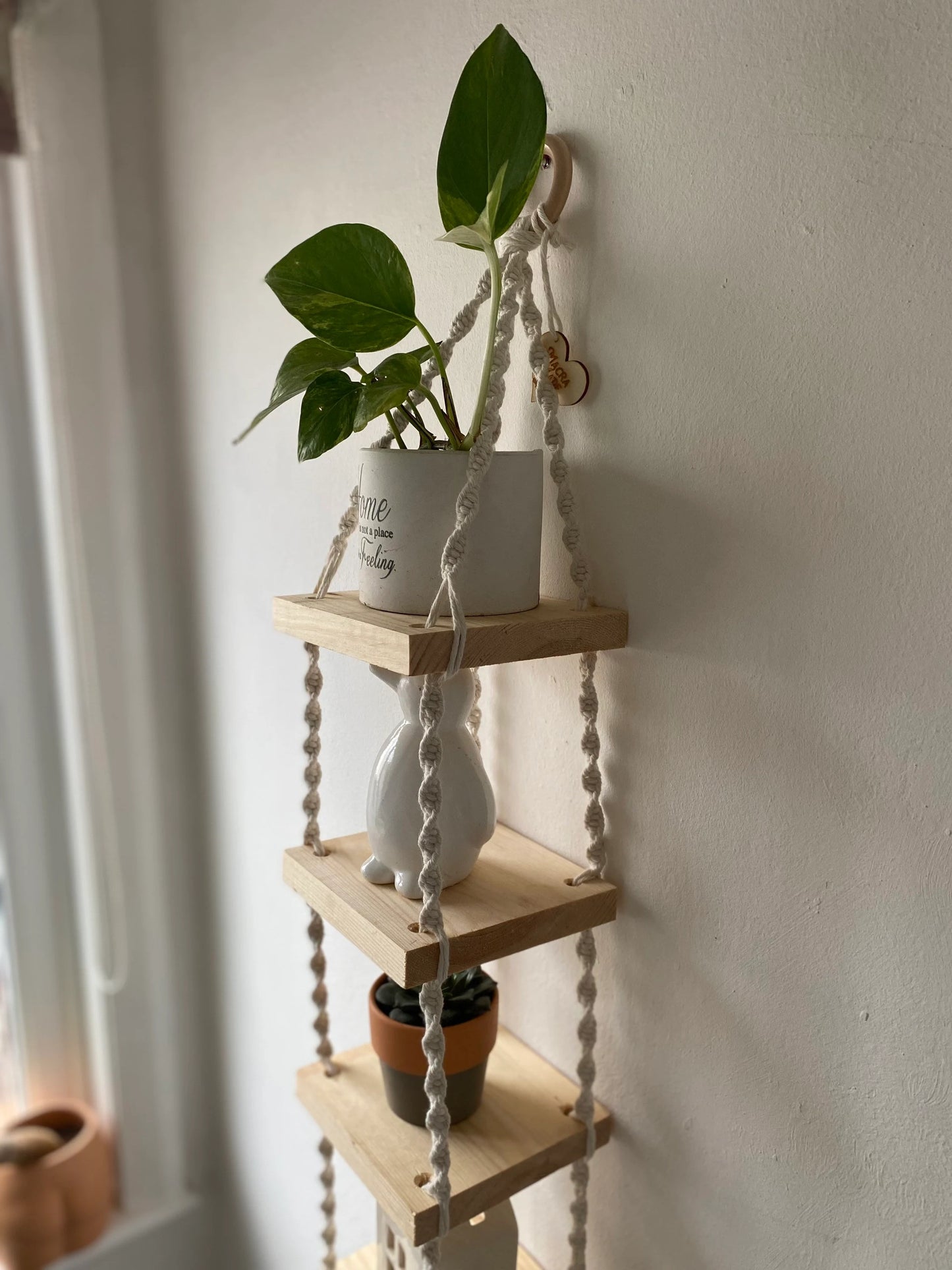 Handmade macrame rope shelves, made with small square wooden shelves, held together with natural knotted rope. Displayed hanging on a white wall, with small houseplants on ornaments displayed on it.