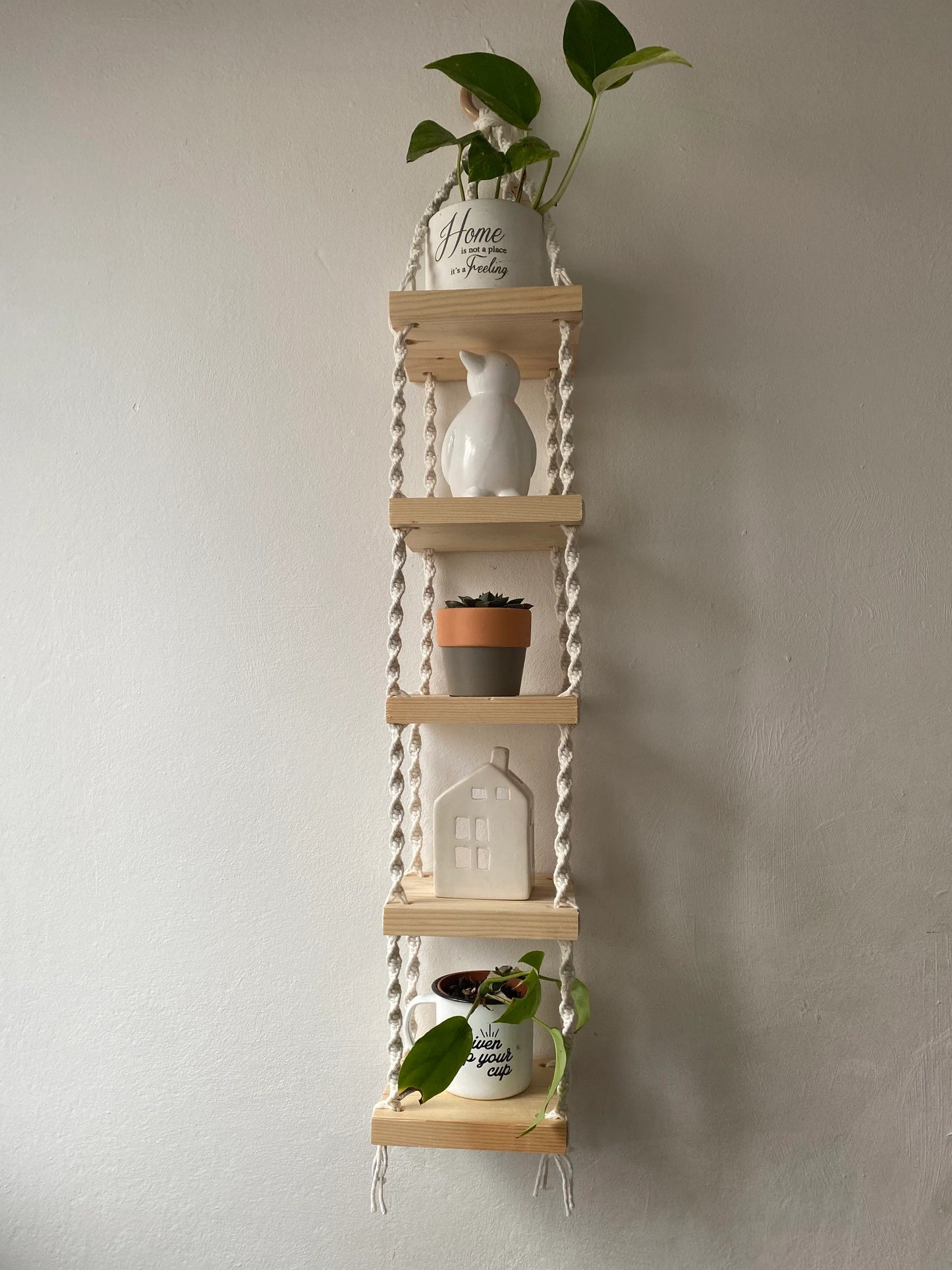 Handmade macrame rope shelves, made with small square wooden shelves, held together with natural knotted rope. Displayed hanging on a white wall, with small houseplants on ornaments displayed on it.