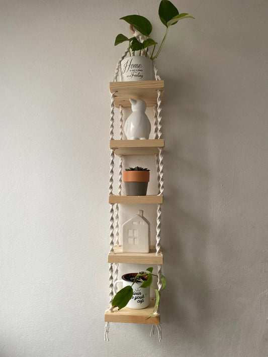 Handmade macrame rope shelves, made with small square wooden shelves, held together with natural knotted rope. Displayed hanging on a white wall, with small houseplants on ornaments displayed on it.