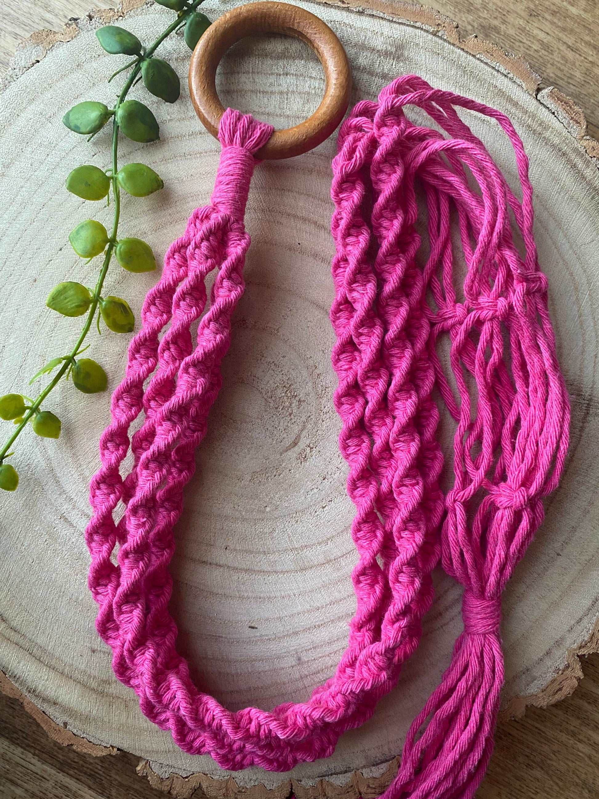A handmade macrame plant hanger, with intricate twisted knot detail, made with bright fuchsia pink recycled cotton and a wooden hanging ring, is coiled on a wooden surface with some green plant vines.