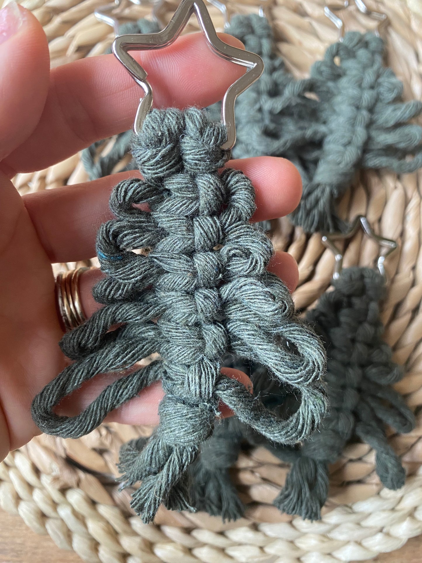 A hand is holding a green Knotted Yarn Hanging Christmas Tree Decoration by Macra-Made-With-Love, crafted from recycled cotton yarns and topped with a star. The background is a woven mat adorned with similar eco-friendly holiday decor. The ornament features intricate knot work and looped fringe at the bottom.