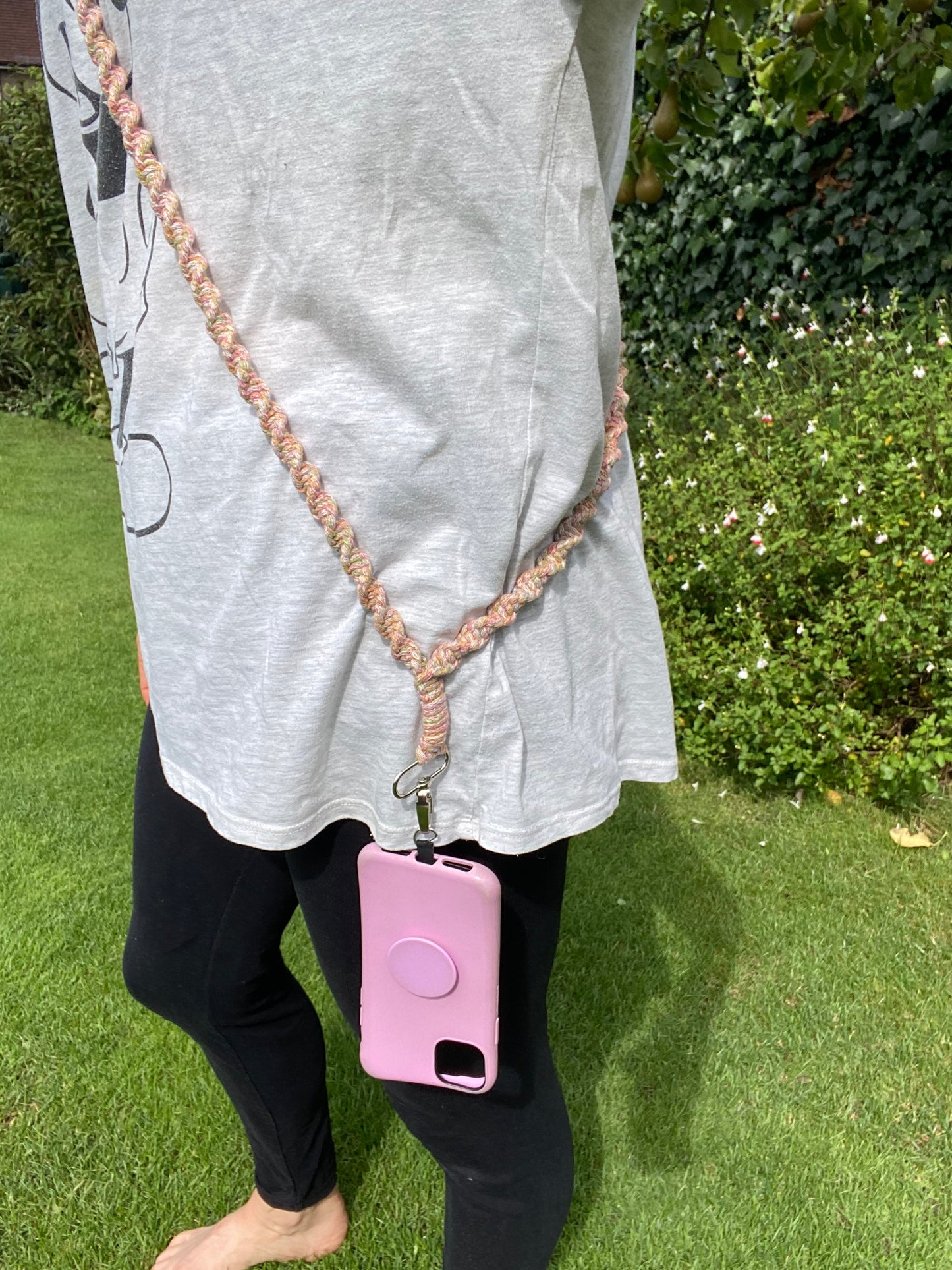 A person stands on a grass lawn, wearing black leggings and a gray shirt with a graphic design. They have an eco-friendly pink phone case attached to a Macra-Made-With-Love Recycled cotton crossbody phone strap slung over their shoulder. Lush green bushes and white flowers are visible in the background.