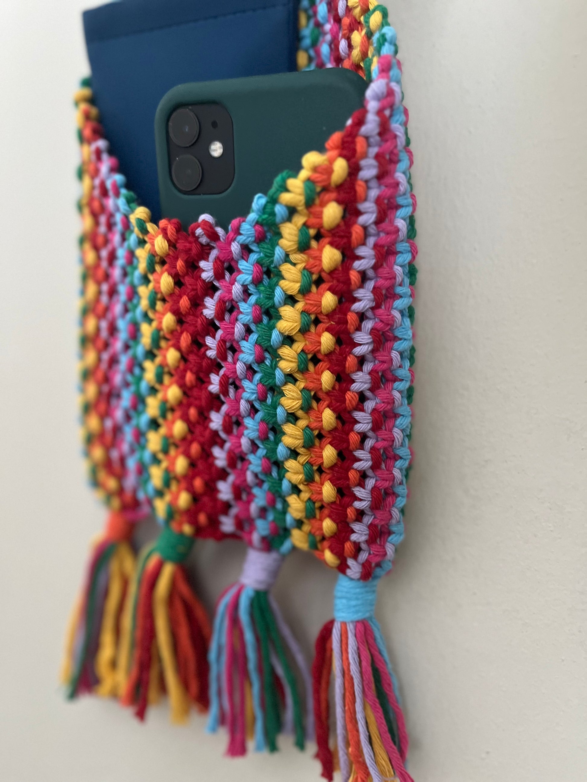 Close-up of a colorful, knitted phone holder hanging on a wall. The **Macra-Made-With-Love Bedside storage pouch book sleeve** features vibrant red, yellow, green, blue, and purple threads and is adorned with five tassels at the bottom. Two smartphones, one green and one blue, are placed inside the holder.