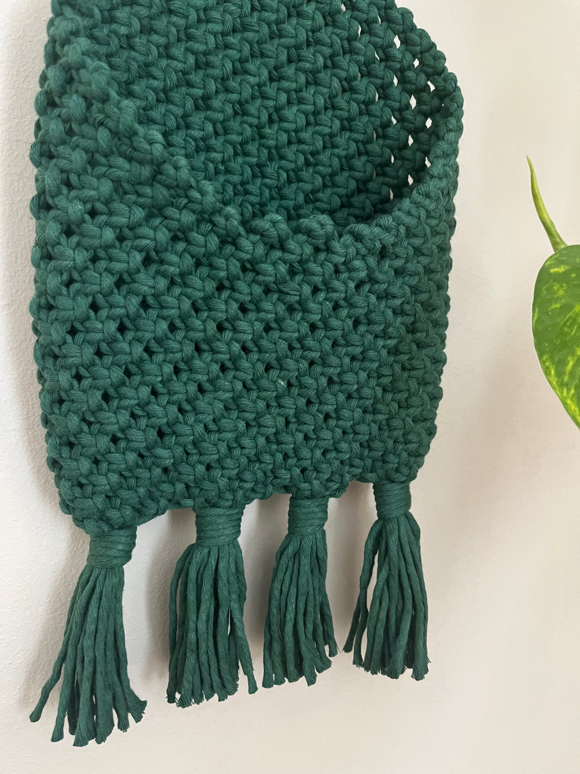 A wall hanging basket made of green woven material, featuring an open pocket and four fringed tassels at the bottom, displayed against a white wall. This Bedside storage pouch book sleeve by Macra-Made-With-Love uses recycled cotton decor. A green plant with heart-shaped leaves is partially visible on the right side of the image.
