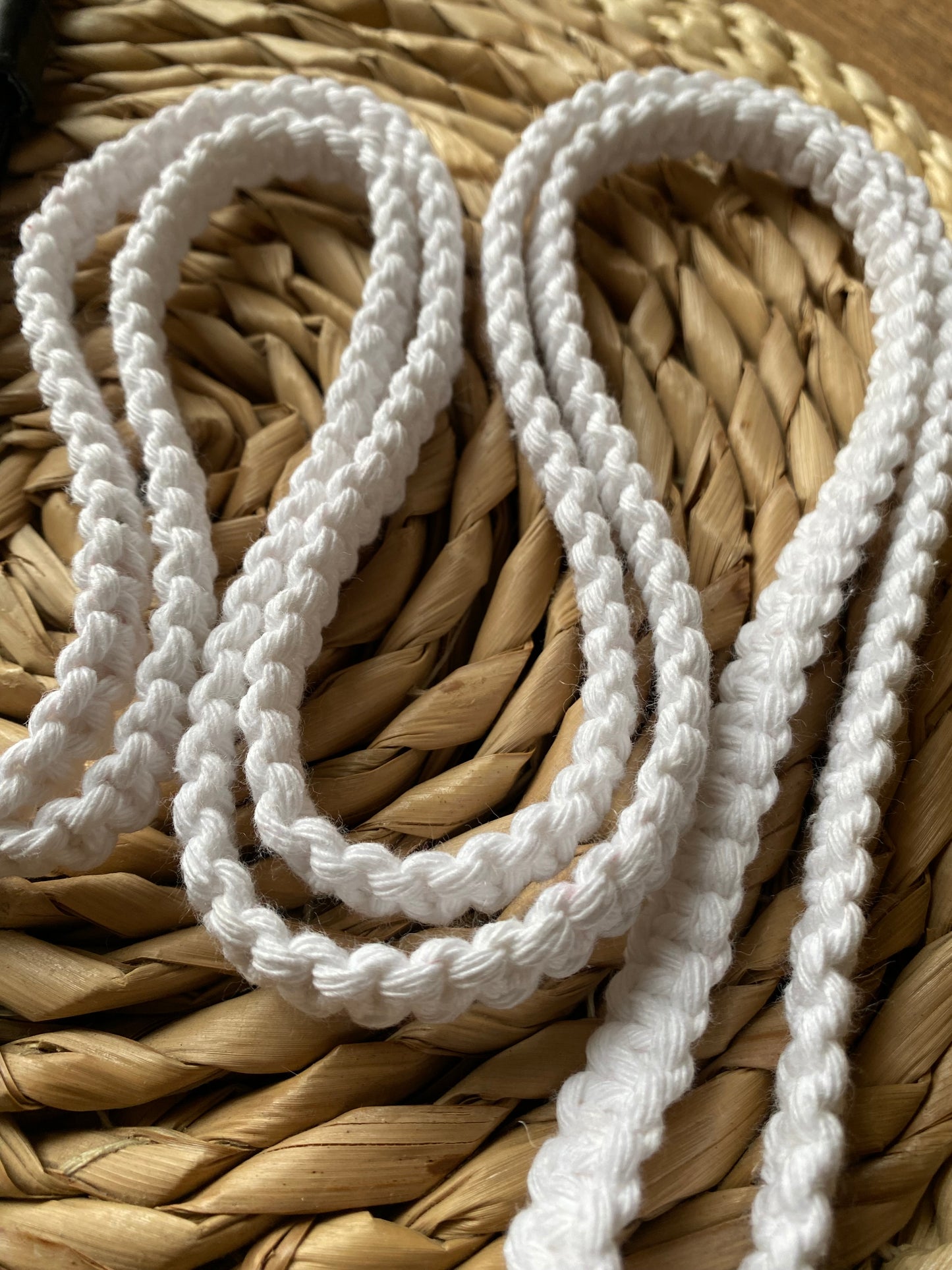 A close-up of white, braided cords arranged in a loose pattern on a woven, natural fiber surface highlights the eco-friendly, handmade Recycled cotton crossbody phone strap from Macra-Made-With-Love. The texture of both the cords and the mat is clearly visible, with the slightly curved cords creating a pleasing visual flow.