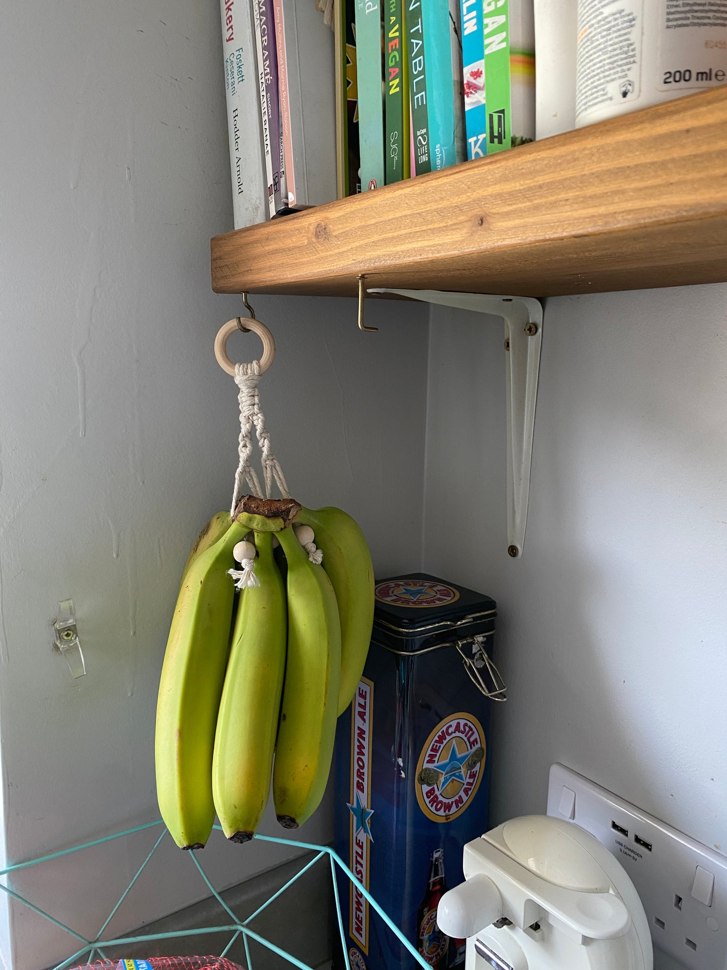 A Kitchen space saving eco friendly banana hanger from Macra-Made-With-Love hangs from a handmade banana holder attached to a wooden shelf. The shelf, supported by white brackets, contains multiple colorful books. Below the shelf, there's a blue, vintage-style tin container and a white electrical outlet on a gray wall, creating an eco-friendly décor.