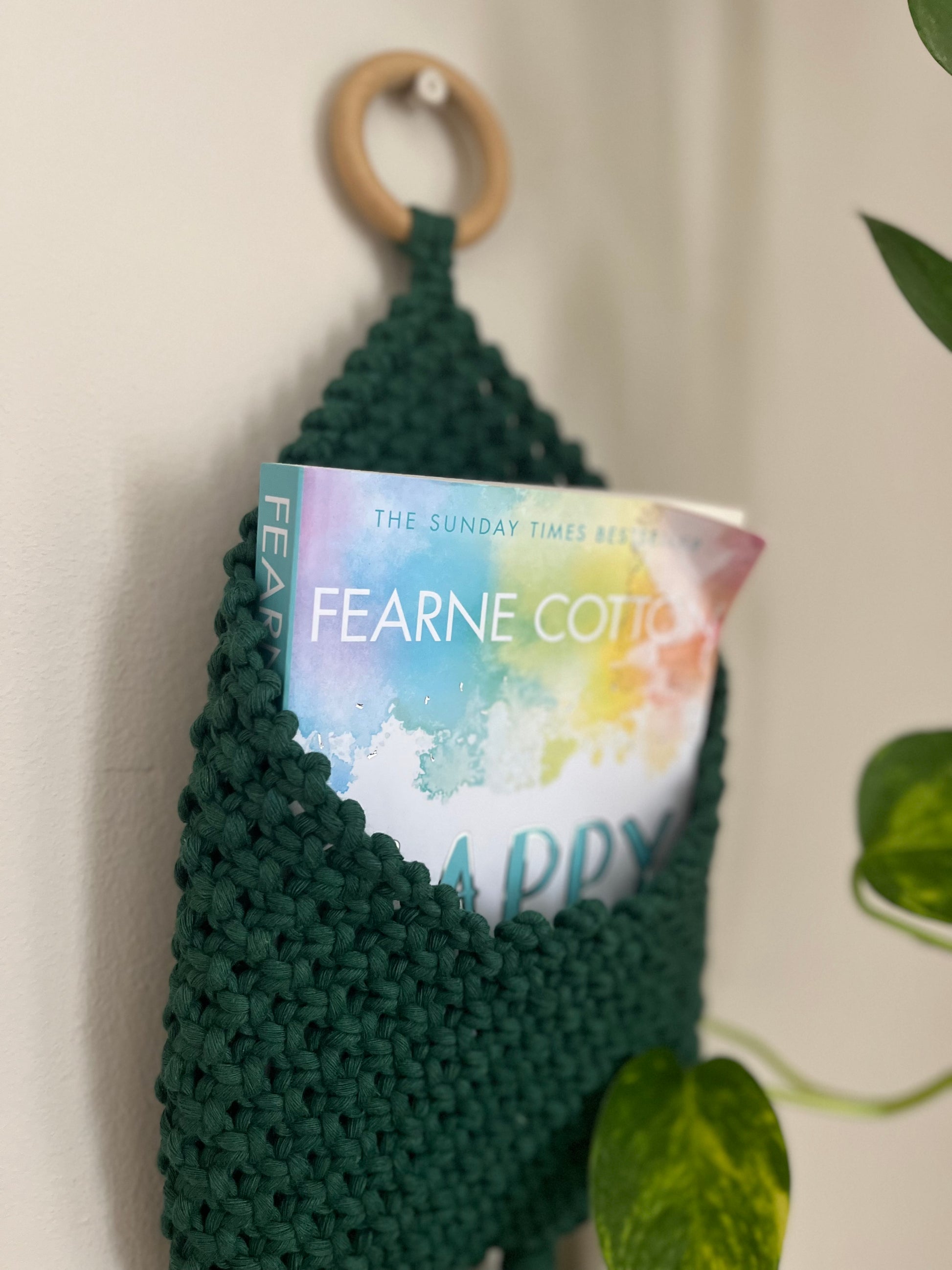 A green crocheted Bedside storage pouch book sleeve by Macra-Made-With-Love, crafted from recycled cotton decor, hangs on a wooden ring against a white wall. Inside the pocket is a book titled "Happy" by Fearne Cotton. A vine plant with heart-shaped leaves is partially visible on the right side of the image, adding an eco-friendly touch.