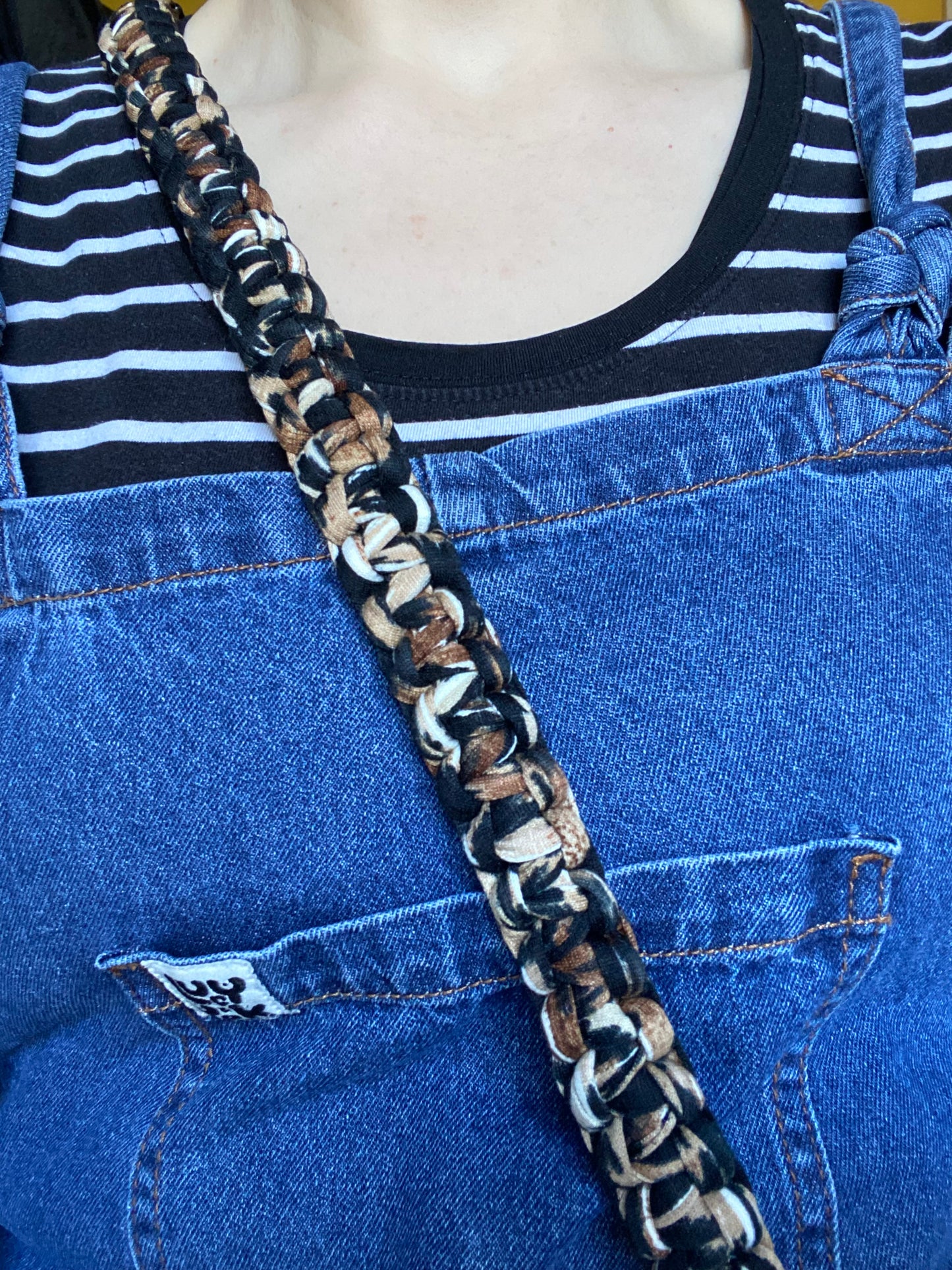 Close-up of a person wearing a black and white striped shirt under blue denim overalls. A handmade Macra-Made-With-Love Recycled cotton crossbody phone strap, featuring an eco-friendly black and tan braided pattern, crosses their chest from the right shoulder to the left side of the overalls.