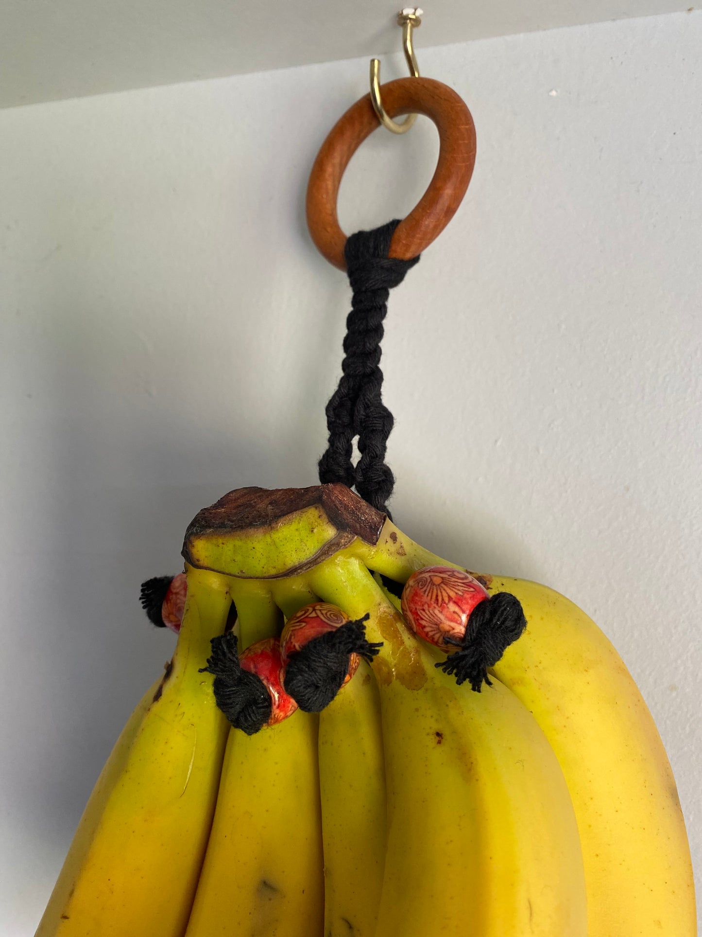 A Macra-Made-With-Love Banana hanger is hanging from a handmade banana holder on the wall. The bananas are tied together with a thick black cord, looped through a wooden ring, and secured to the hook. Small red and black tassels add charm to this eco-friendly home decor piece.